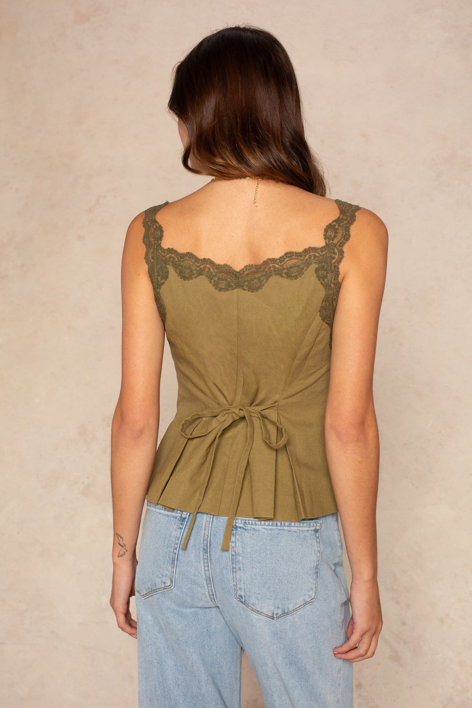 A woman with long brown hair, seen from behind, wears the Georgie Tillie Top—an olive green sleeveless blouse with lace trim and adjustable back tie—paired with light blue high-waisted jeans, against a neutral background.
