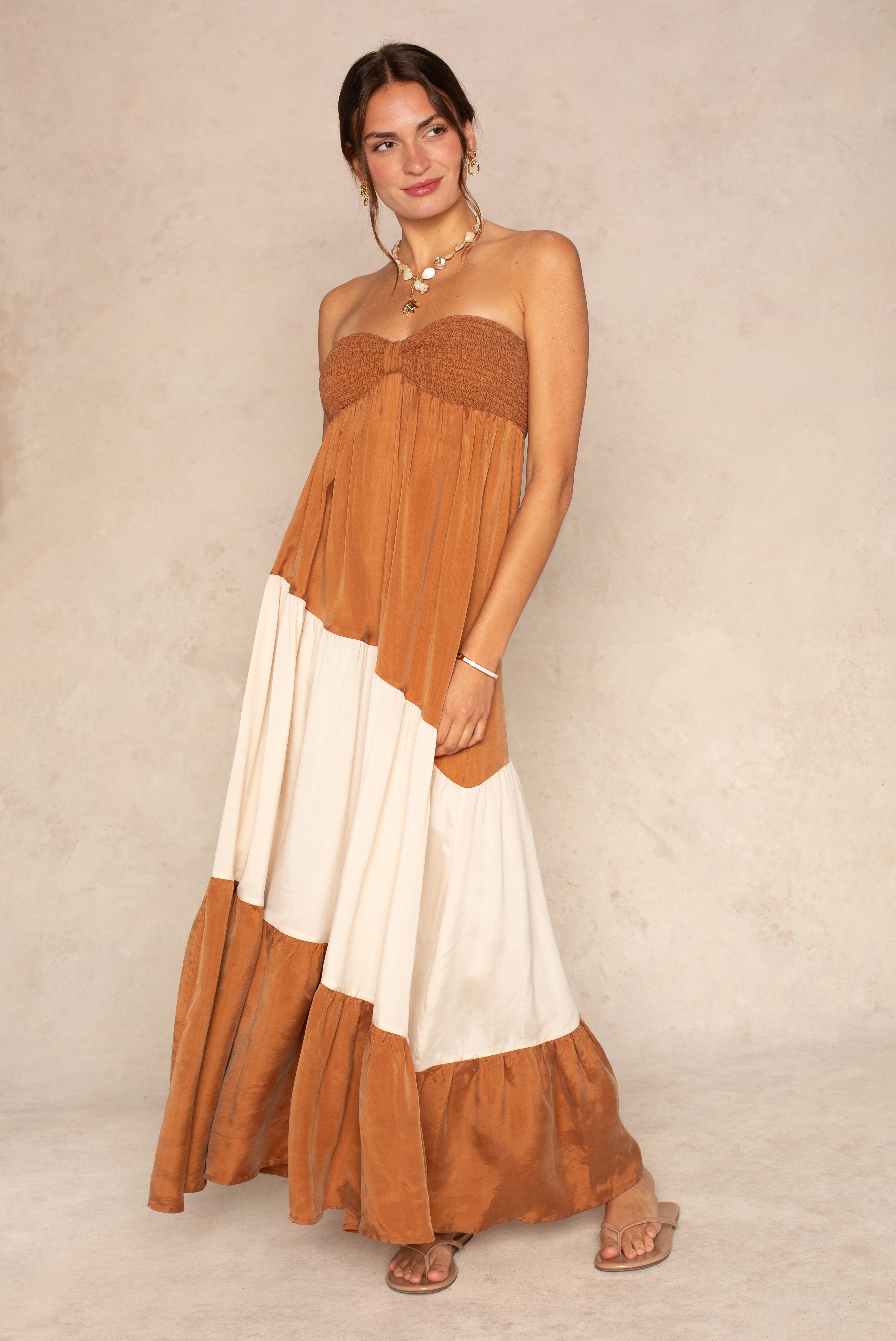 A woman models The Sashia Maxi Dress by Sancia in Brown Multi, standing against a neutral background with sandals and a relaxed smile—an effortlessly chic vacation look.