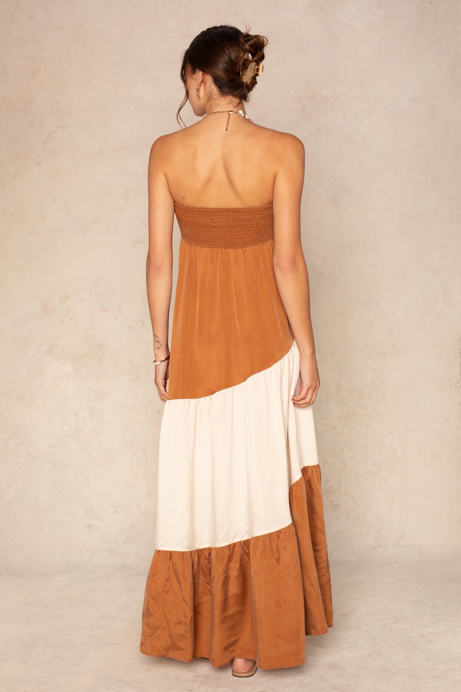 A woman faces away, wearing The Sashia Maxi Dress by Sancia—a strapless colorblock maxi in brown and cream, styled with a loose bun against a neutral textured background. Perfect as a vacation dress.