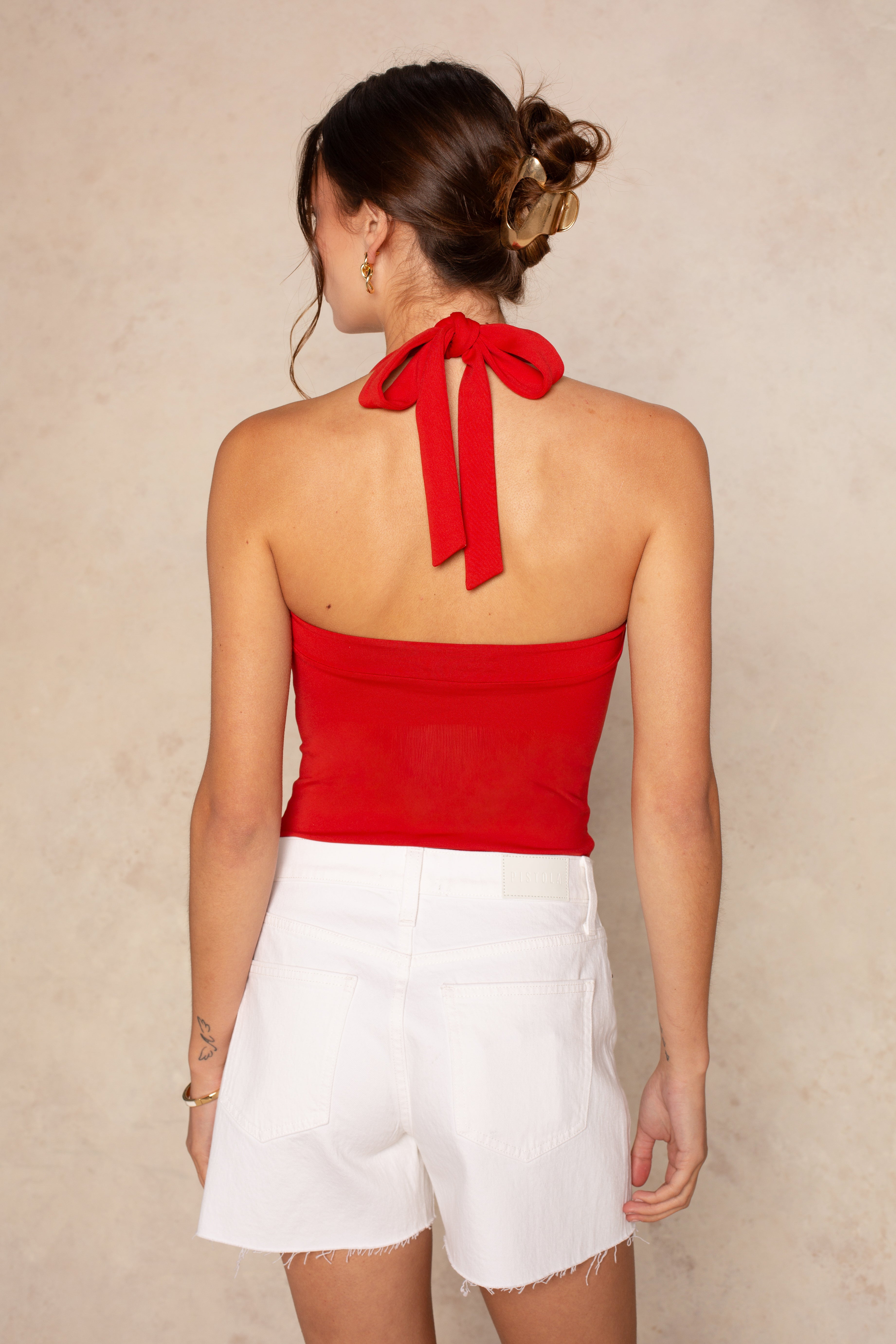 A woman with brown hair in a low bun is seen from behind, showcasing the open back of her Georgie Whitney Compression Tie Halter in red, paired with high-waisted white shorts against a neutral, textured wall.