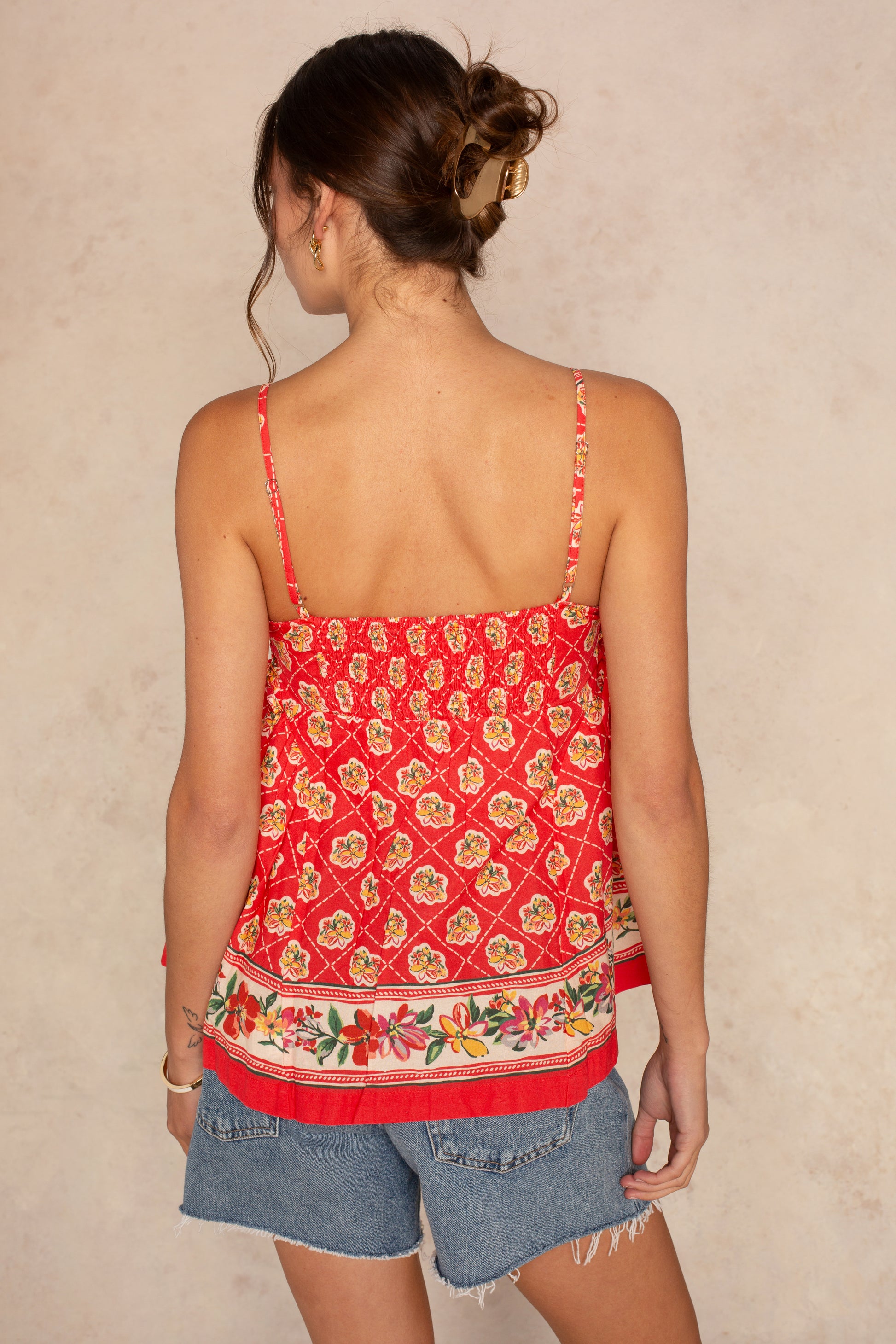 A woman with her hair in a loose bun faces away, wearing the Isla Lila Floral Peplum Cami Top and blue denim shorts against a light beige background.