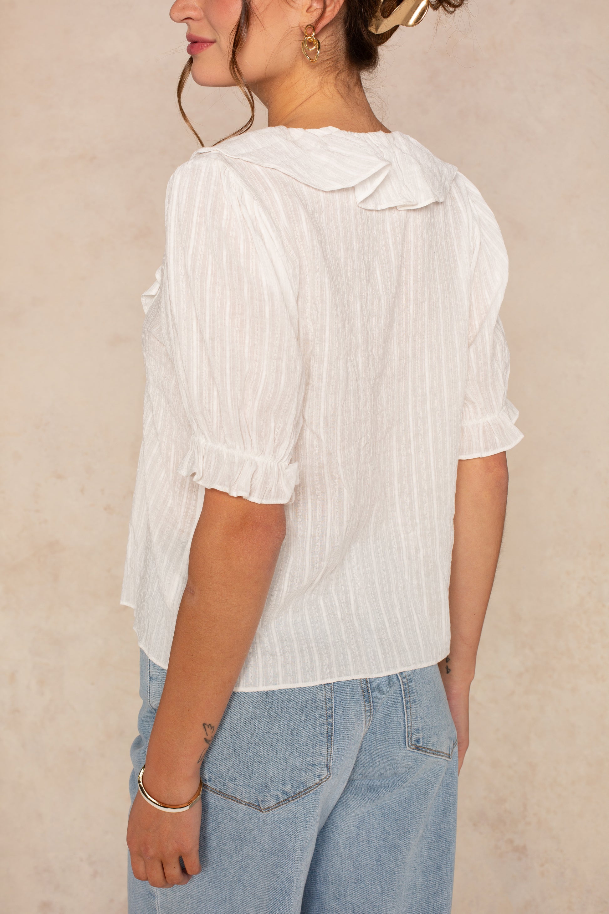 A woman wears the Georgie Aimee Ruffle Short Sleeve Blouse in white, featuring textured fabric, ruffled sleeves and collar, paired with light blue jeans. She’s shown from the back and side with her hair up and gold earrings visible.
