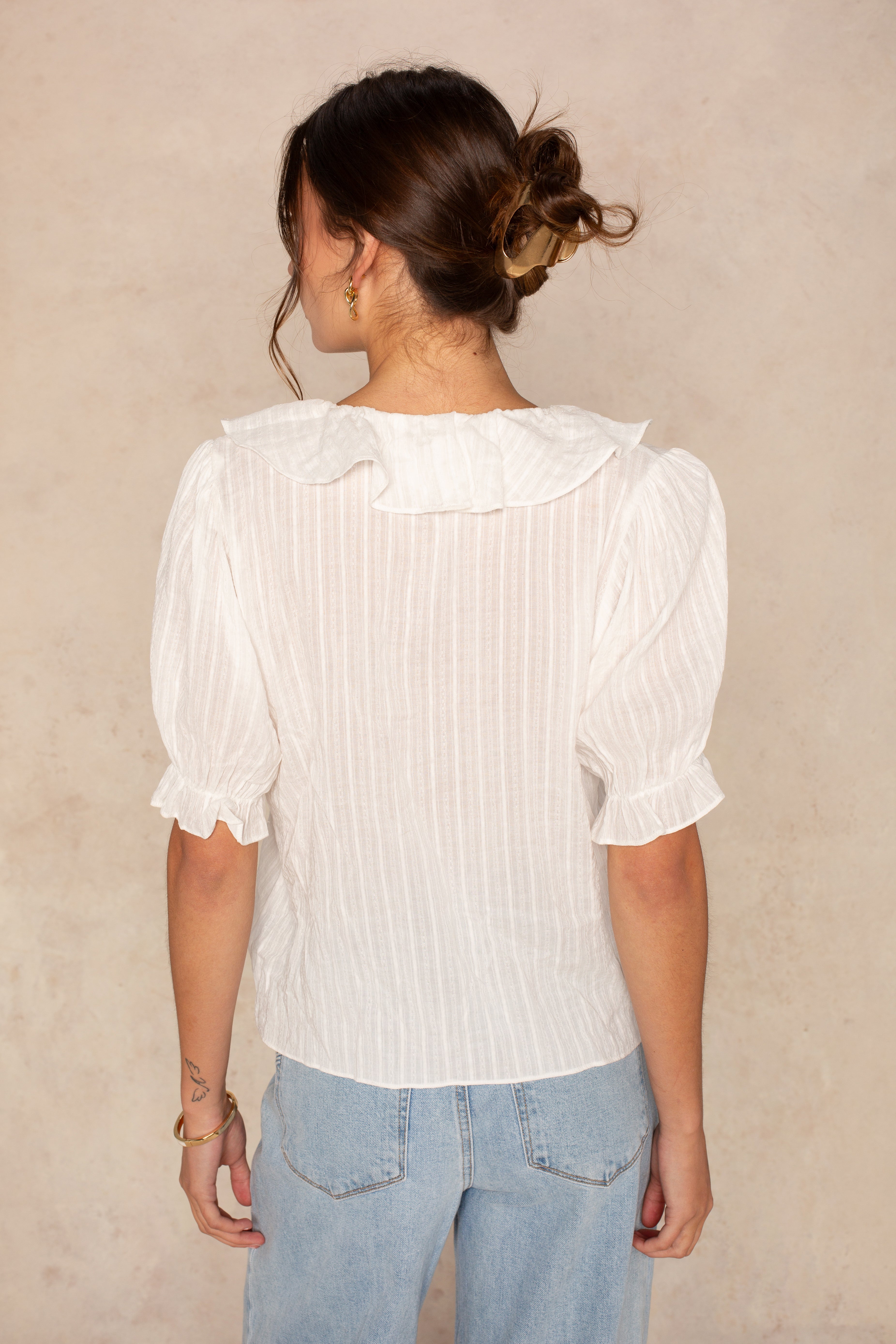 A woman with brown hair in a messy bun faces away, wearing the Georgie Aimee Ruffle Short Sleeve Blouse and light blue jeans against a textured beige background.