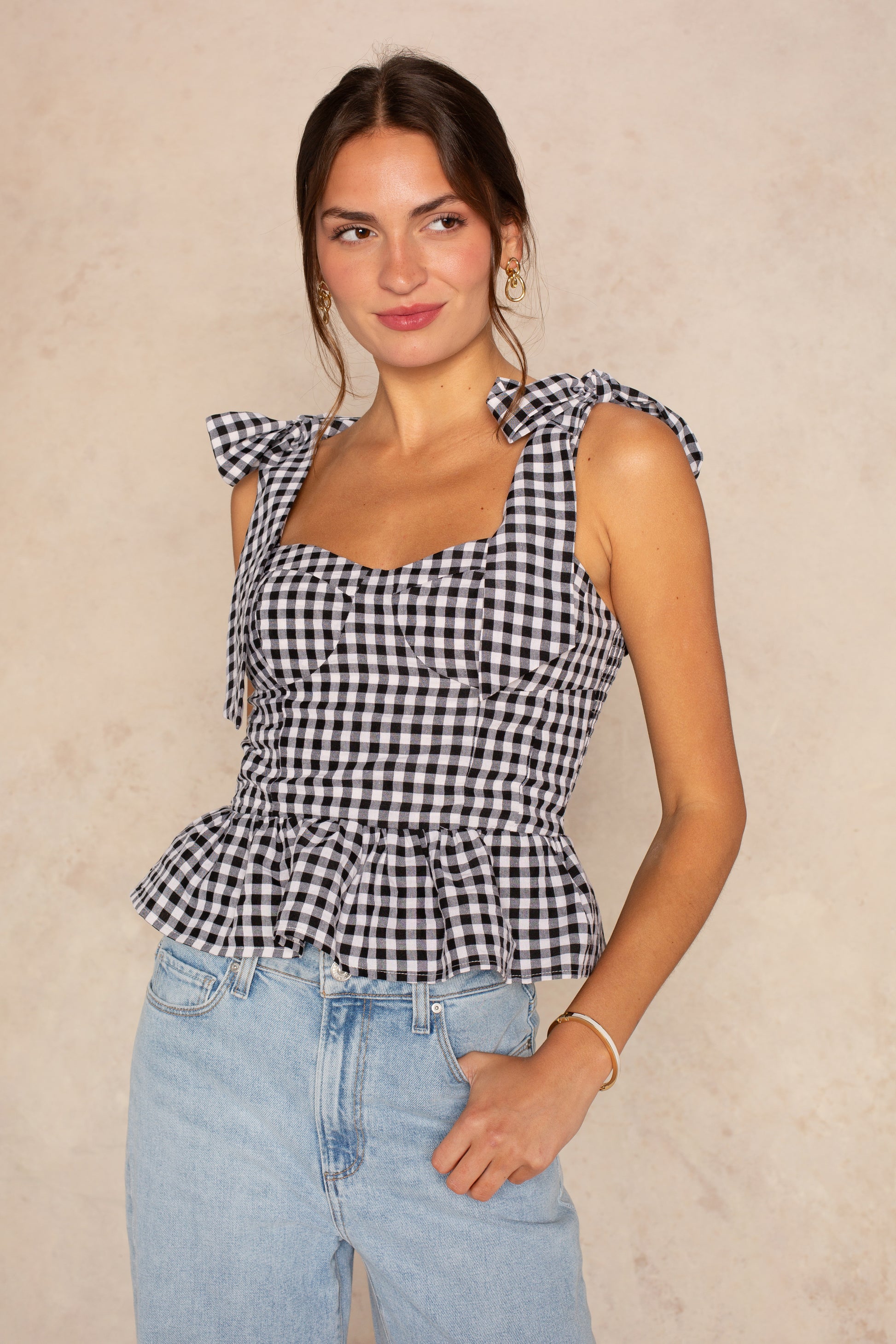 A woman wears the Isla Elsie Bow Shoulder Peplum Tank in Black Multi with tied straps and peplum hem, paired with light blue jeans, standing against a light beige background.