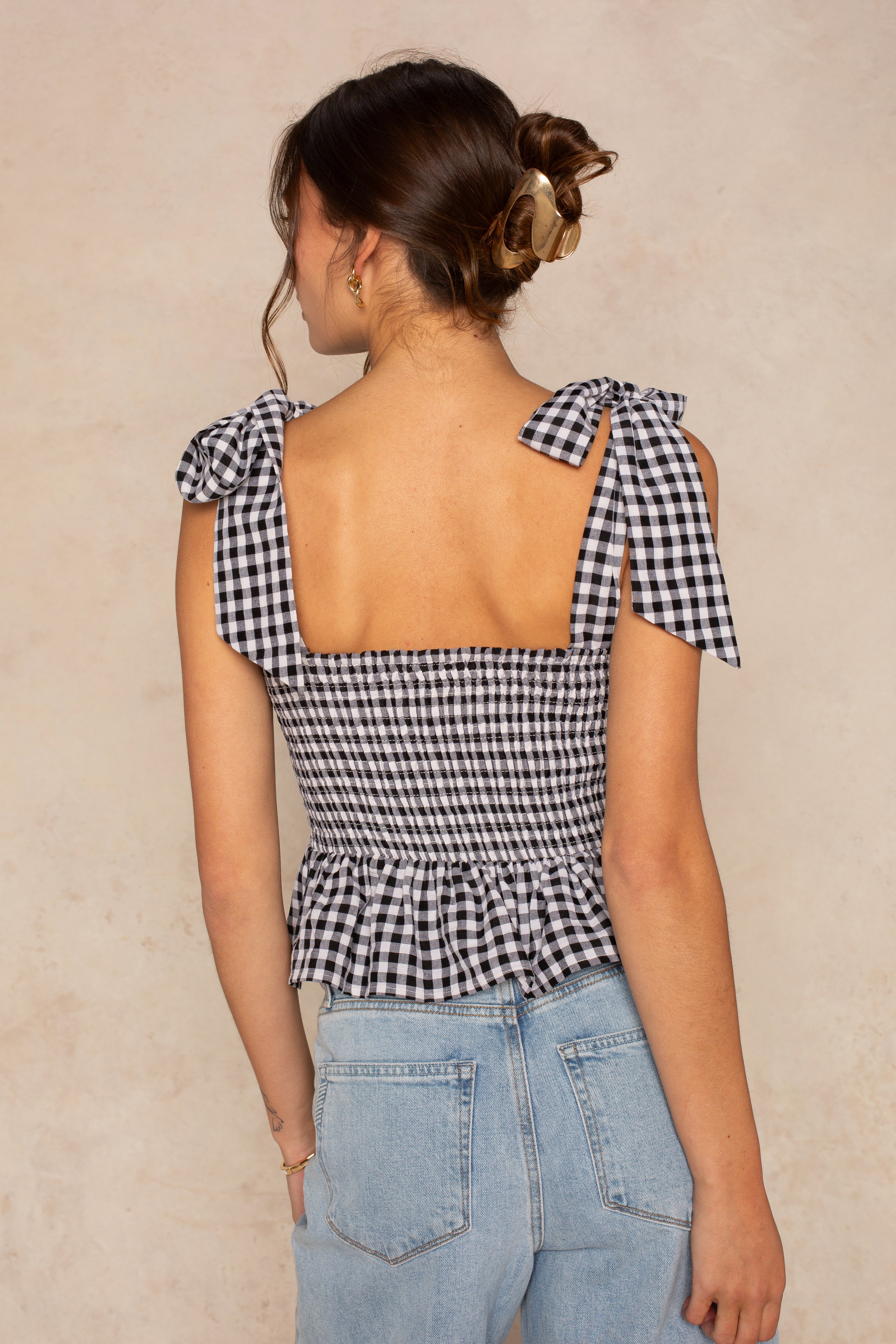 A woman with brown hair in a claw clip wears the Isla Elsie Bow Shoulder Peplum Tank—a black-and-white gingham sleeveless top with bow-tied straps and ruffled hem—paired with light blue high-waisted jeans, against a beige background.