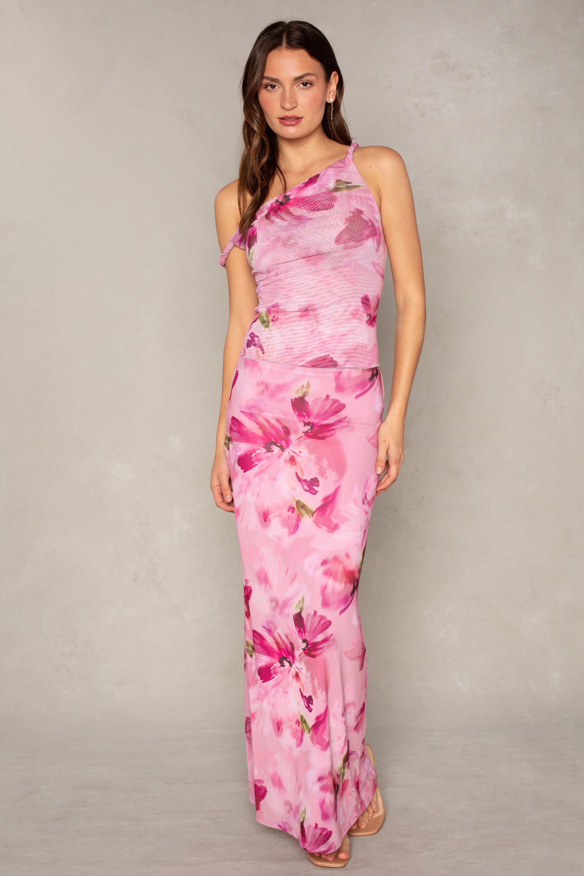 A woman with long, wavy brown hair and a neutral expression stands against a neutral background wearing the Isla Flora Mesh Twist Strap Top, featuring a one-shoulder design and floral pattern.