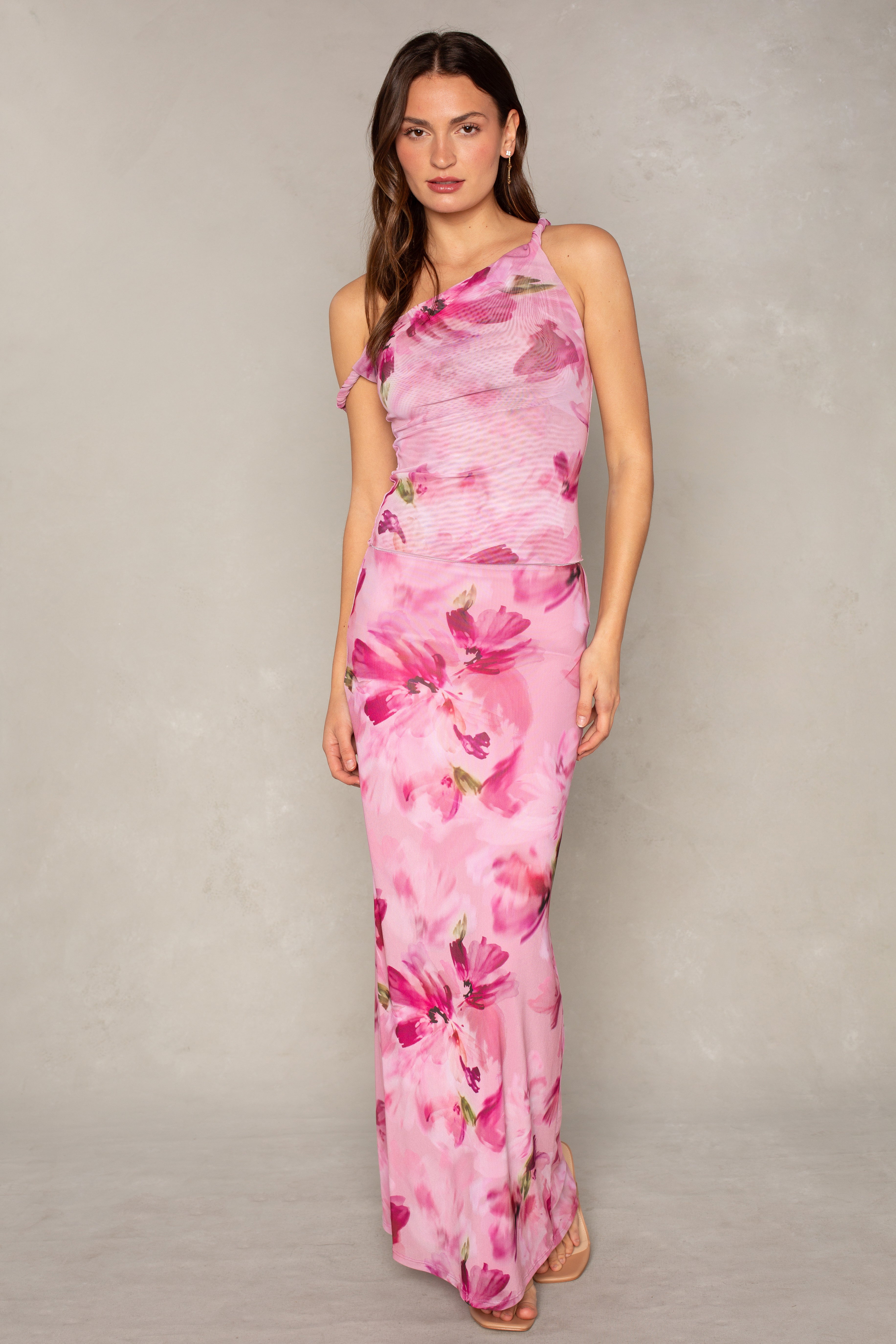 A woman with long, wavy brown hair and a neutral expression stands against a neutral background wearing the Isla Flora Mesh Twist Strap Top, featuring a one-shoulder design and floral pattern.