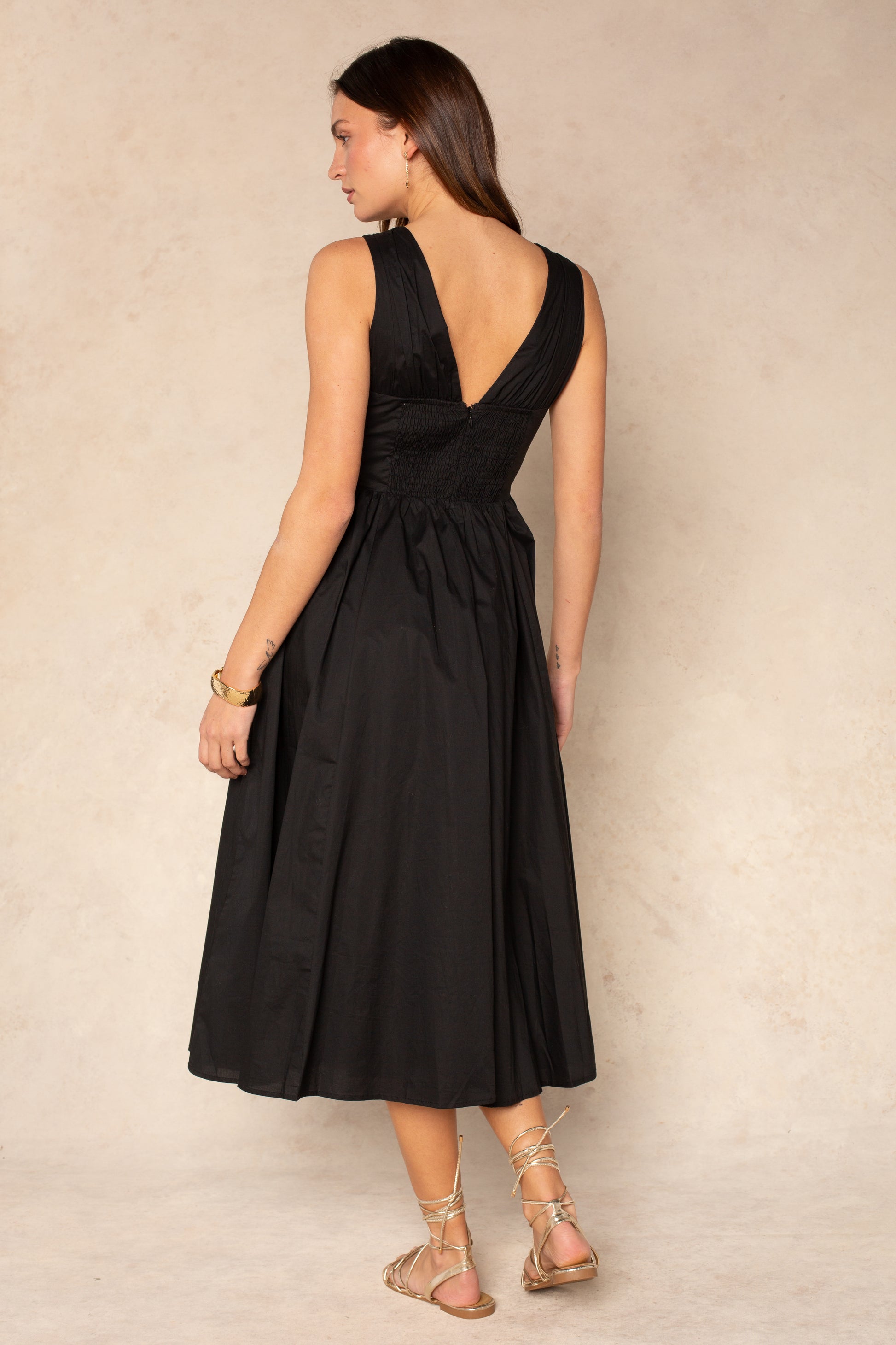 A woman stands on a neutral background wearing the Georgie Vivianne Corset Midi Dress—a sleeveless black dress with a fitted bodice, deep V back, gathered waist, and knee-length skirt—styled with tan lace-up sandals and a bracelet.