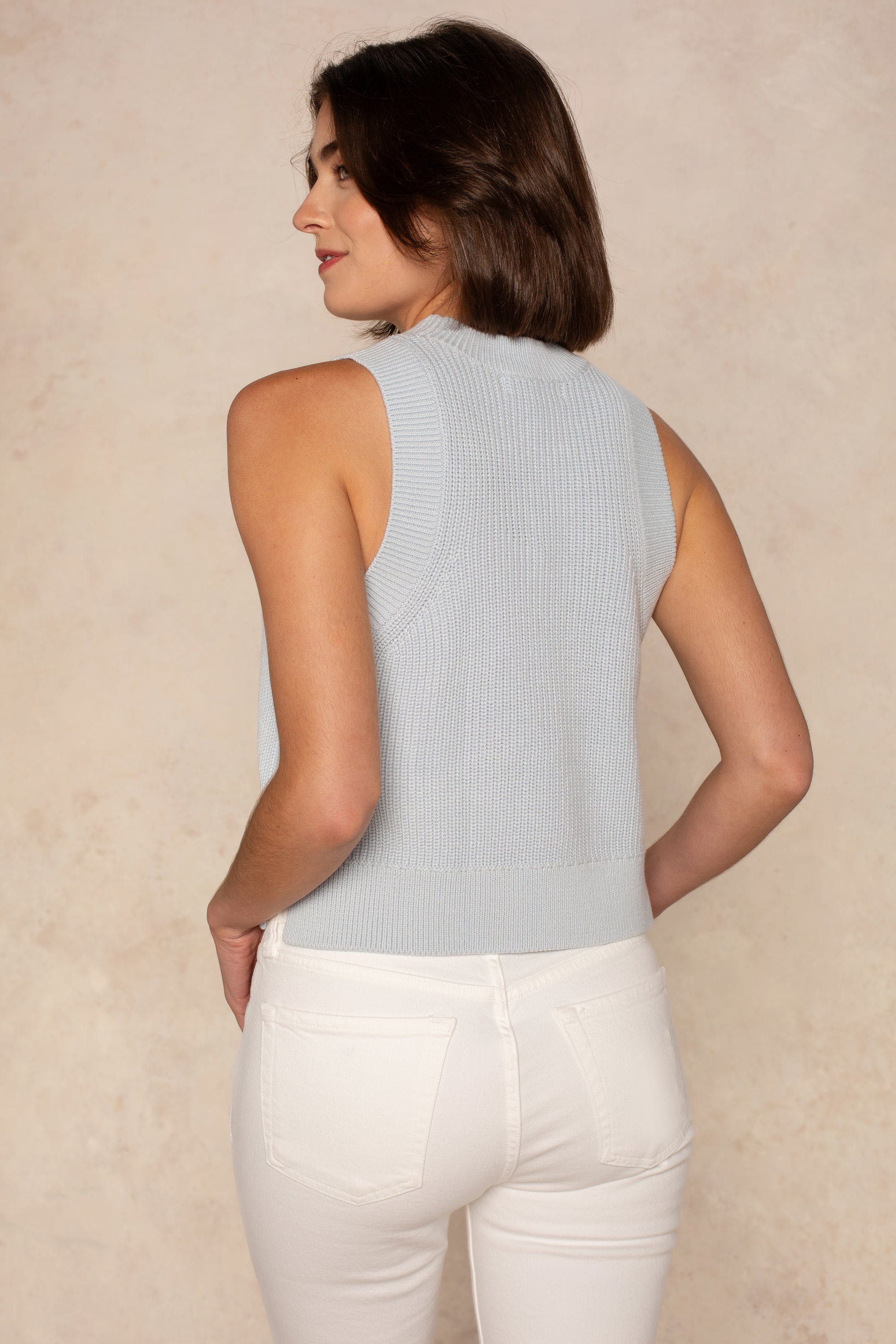 A woman with short brown hair is seen from behind, wearing the Georgie Cleo Sleeveless Crew Sweater in pastel blue and high-waisted white jeans against a light beige background.