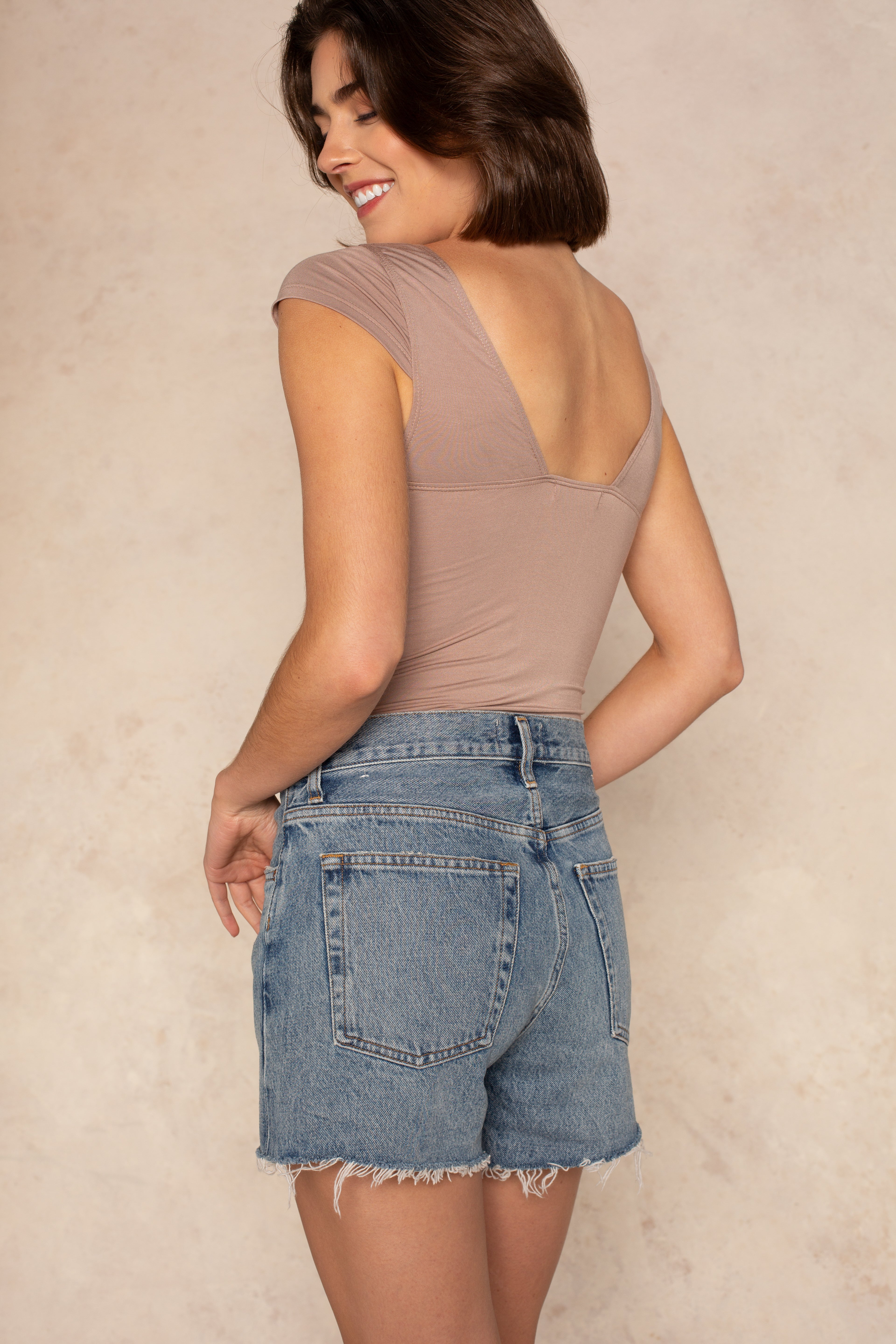 A woman with short brown hair, wearing the Isla Daisy Cap Sleeve Knit Top in taupe and high-waisted denim shorts, stands smiling slightly turned away from the camera against a neutral background.