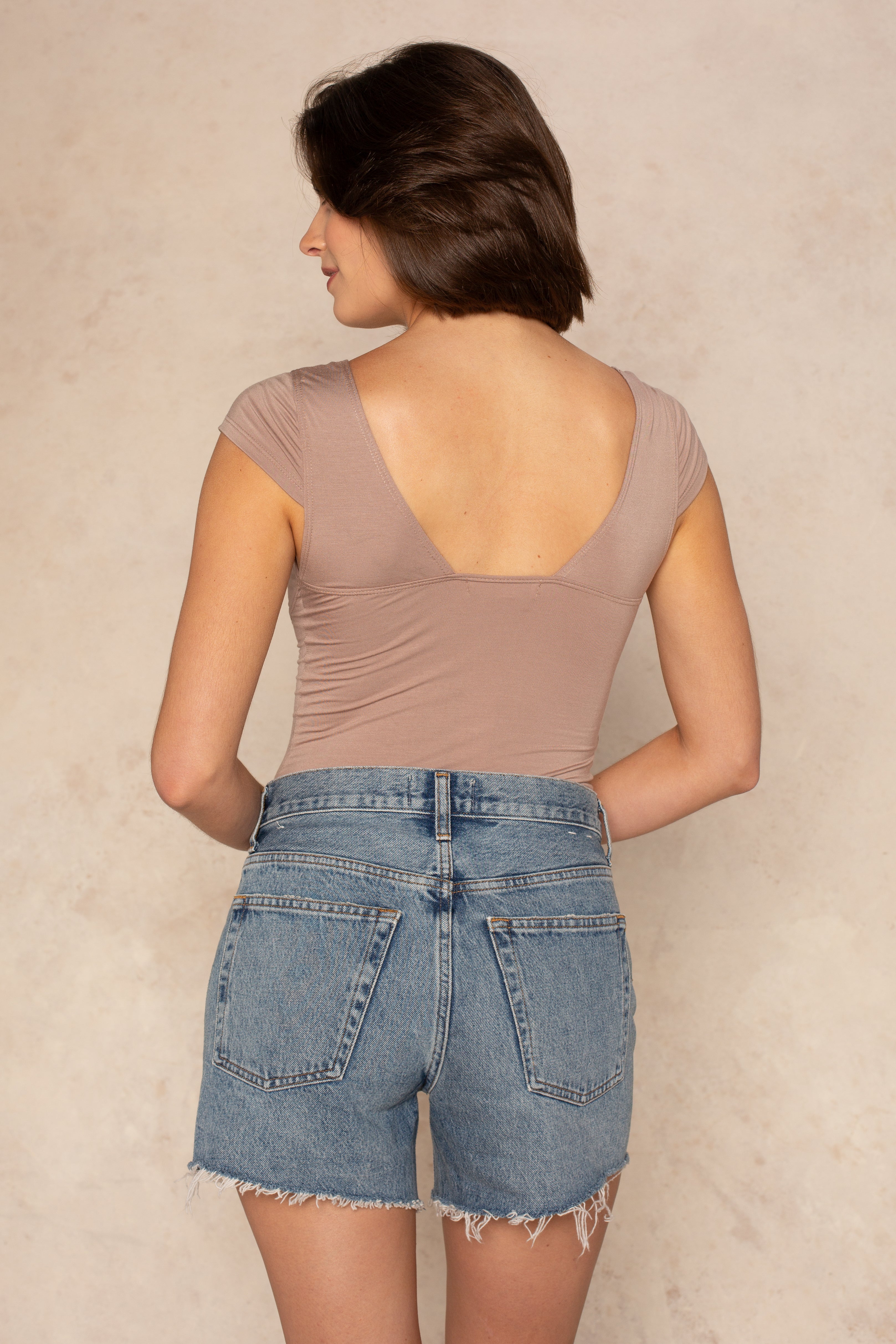 A person with short brown hair is seen from the back, wearing the Isla Daisy Cap Sleeve Knit Top in taupe and high-waisted blue denim shorts with frayed hems. The background is a light, neutral color.