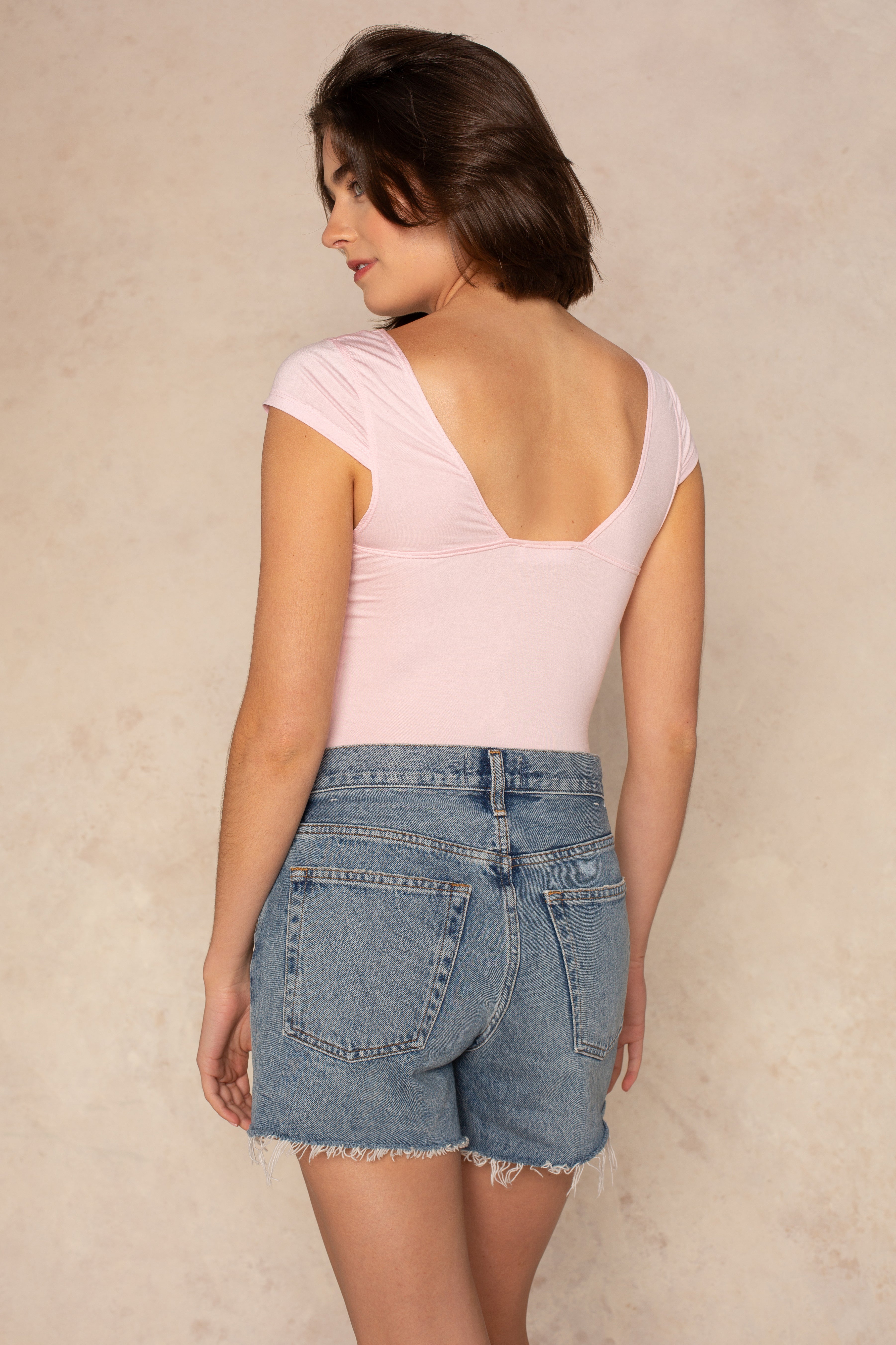 A woman stands facing away, wearing the Isla Daisy Cap Sleeve Knit Top in light pink with a deep V-back and high-waisted, frayed denim shorts. The stretchy fabric flatters her silhouette against a light beige background.