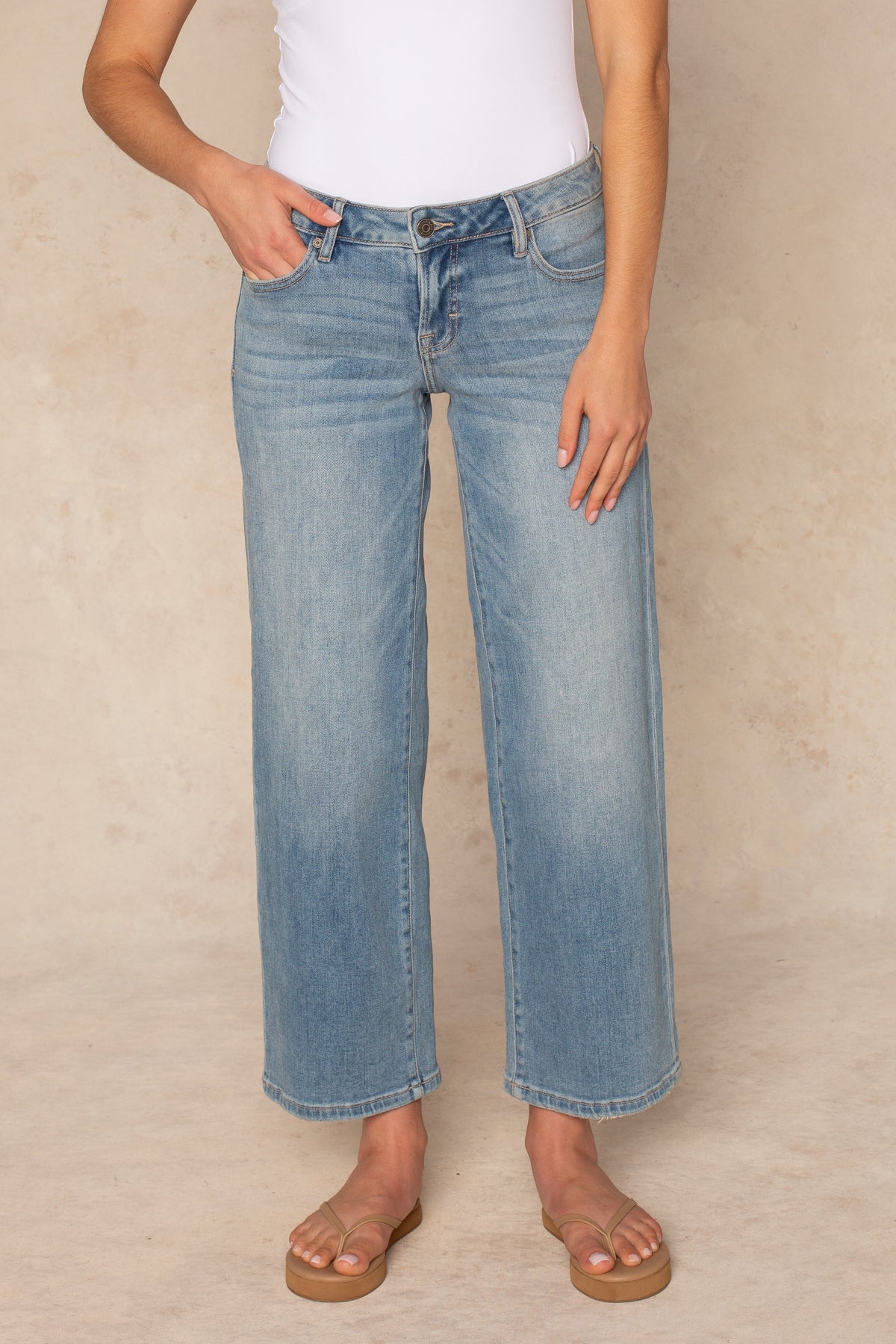 A woman with short brown hair poses against a beige background, wearing a white deep V-neck top, Isla's Lola Mid Rise Straight Leg denim in light blue, and tan flat sandals. She stands relaxed, looking slightly to the side.