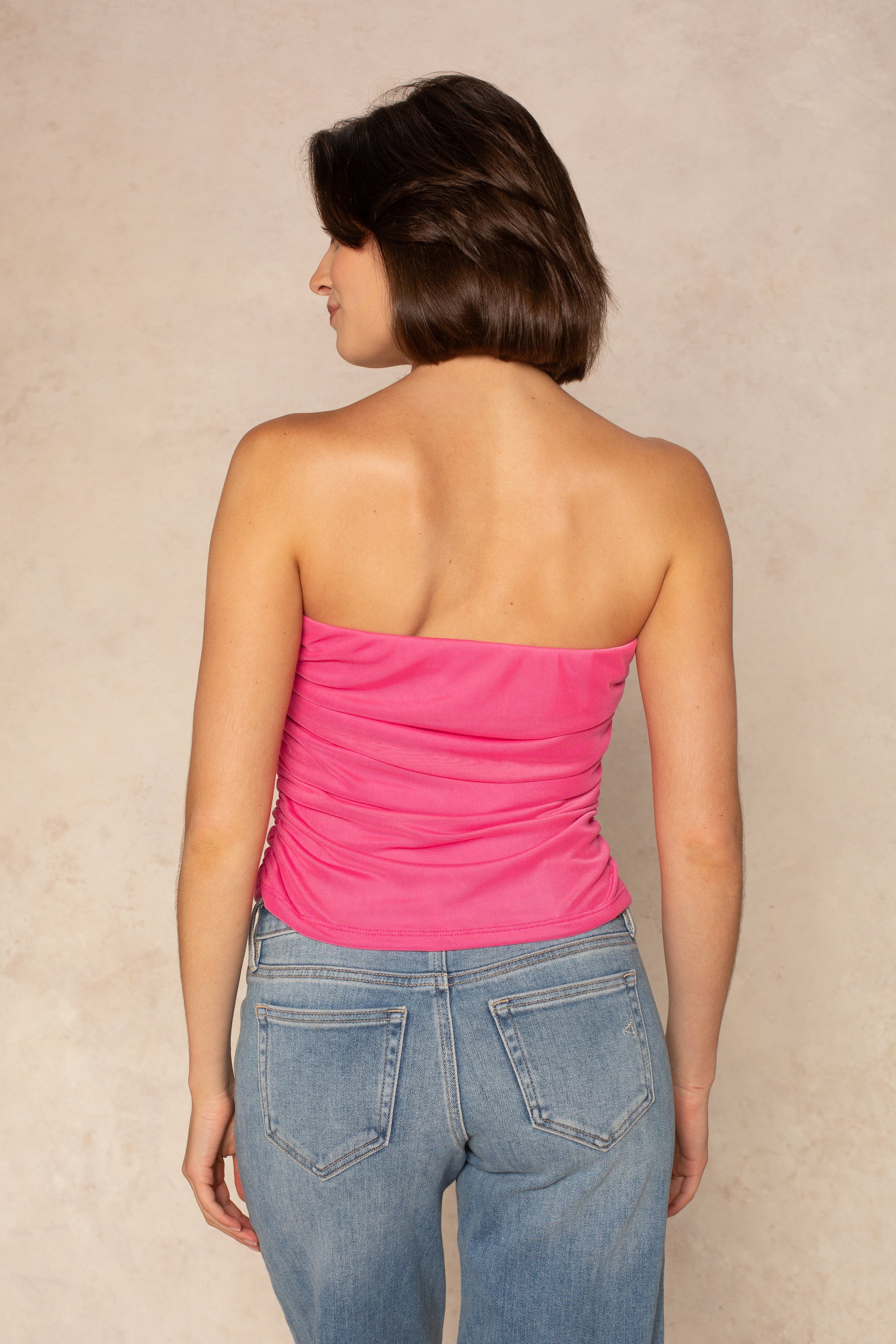 A woman with short brown hair stands with her back to the camera, wearing Isla’s Mia Mesh Tube dress in bright pink and light blue jeans against a neutral beige background.