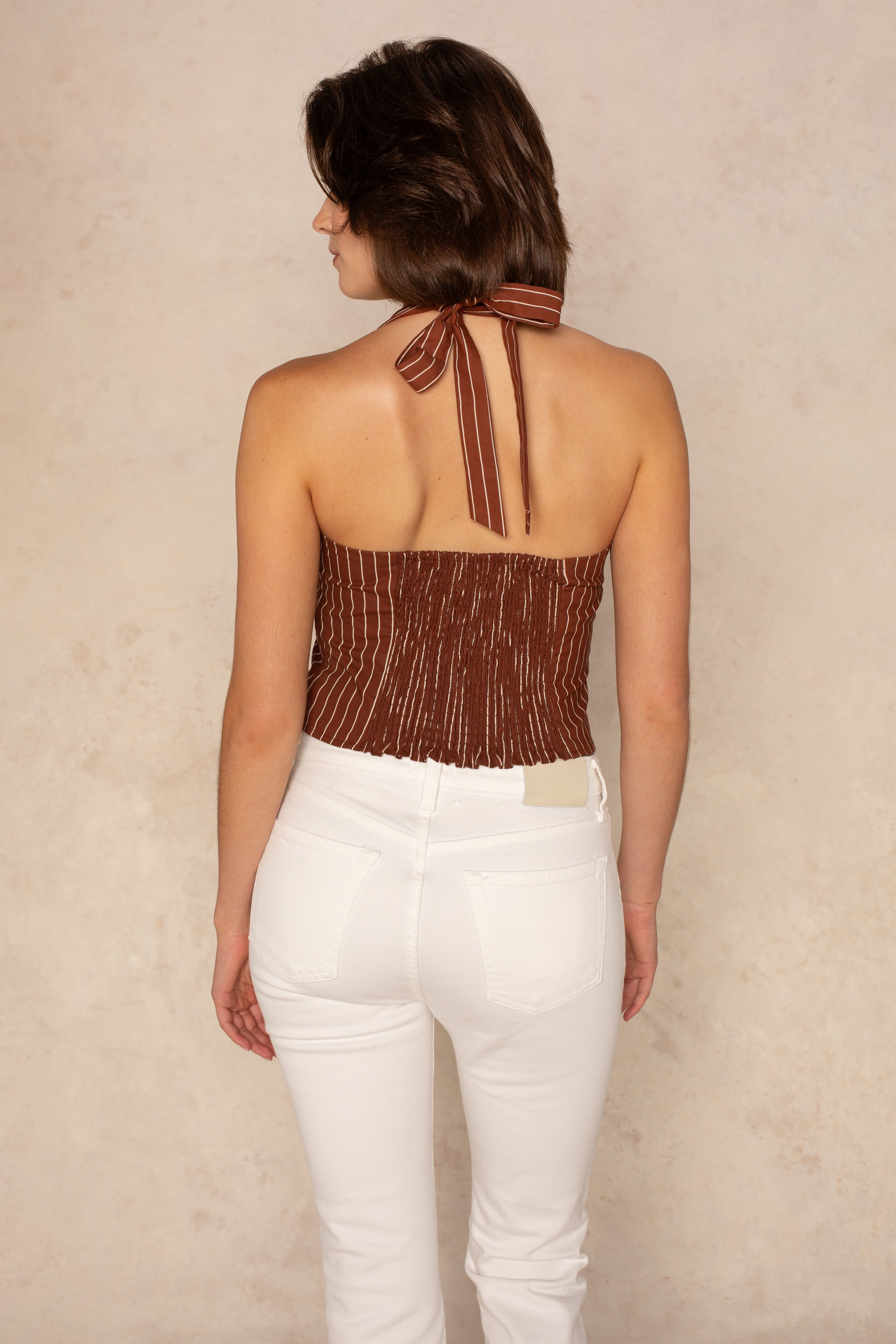 A woman with chin-length dark hair, facing away from the camera, wears the Georgie Harper Halter Linen Top—brown with white stripes—and white high-waisted jeans against a neutral beige background.