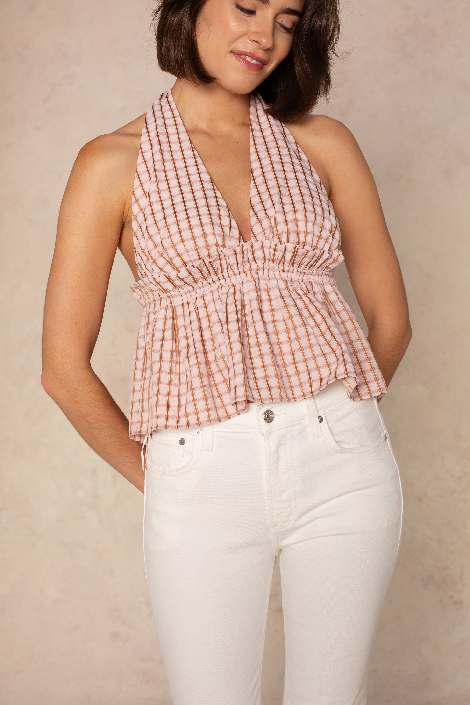 A woman wears the Isla Mira Halter Top in Pink Multi, featuring a smocked empire waist, paired with high-waisted white jeans. She stands against a beige background with a slight smile, eyes closed, and hands behind her back.