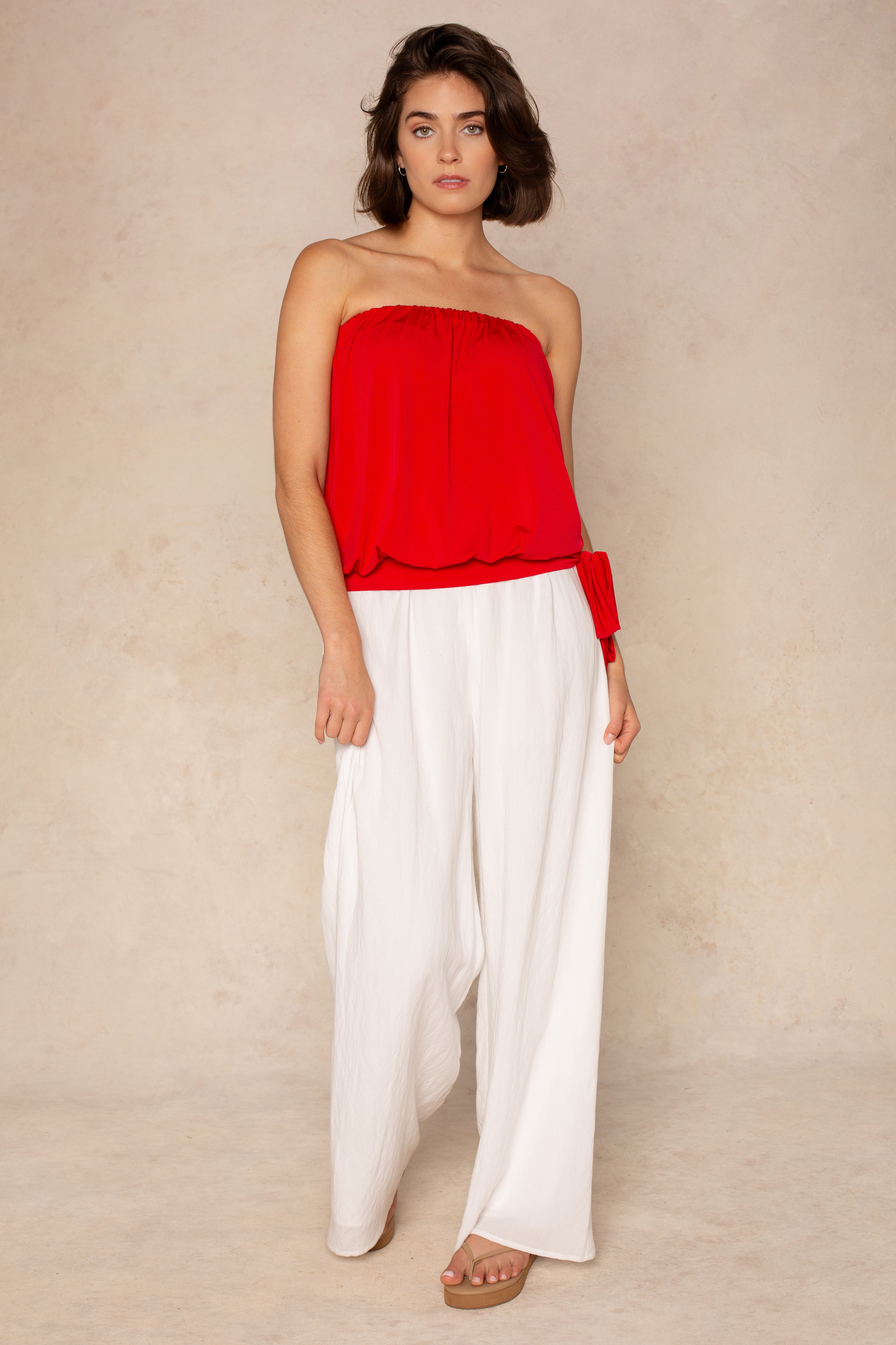 A woman with short, wavy brown hair, barefoot and relaxed against a neutral background, wears the Georgie Gemma Side Tie Tube Top in red paired with loose white pants.
