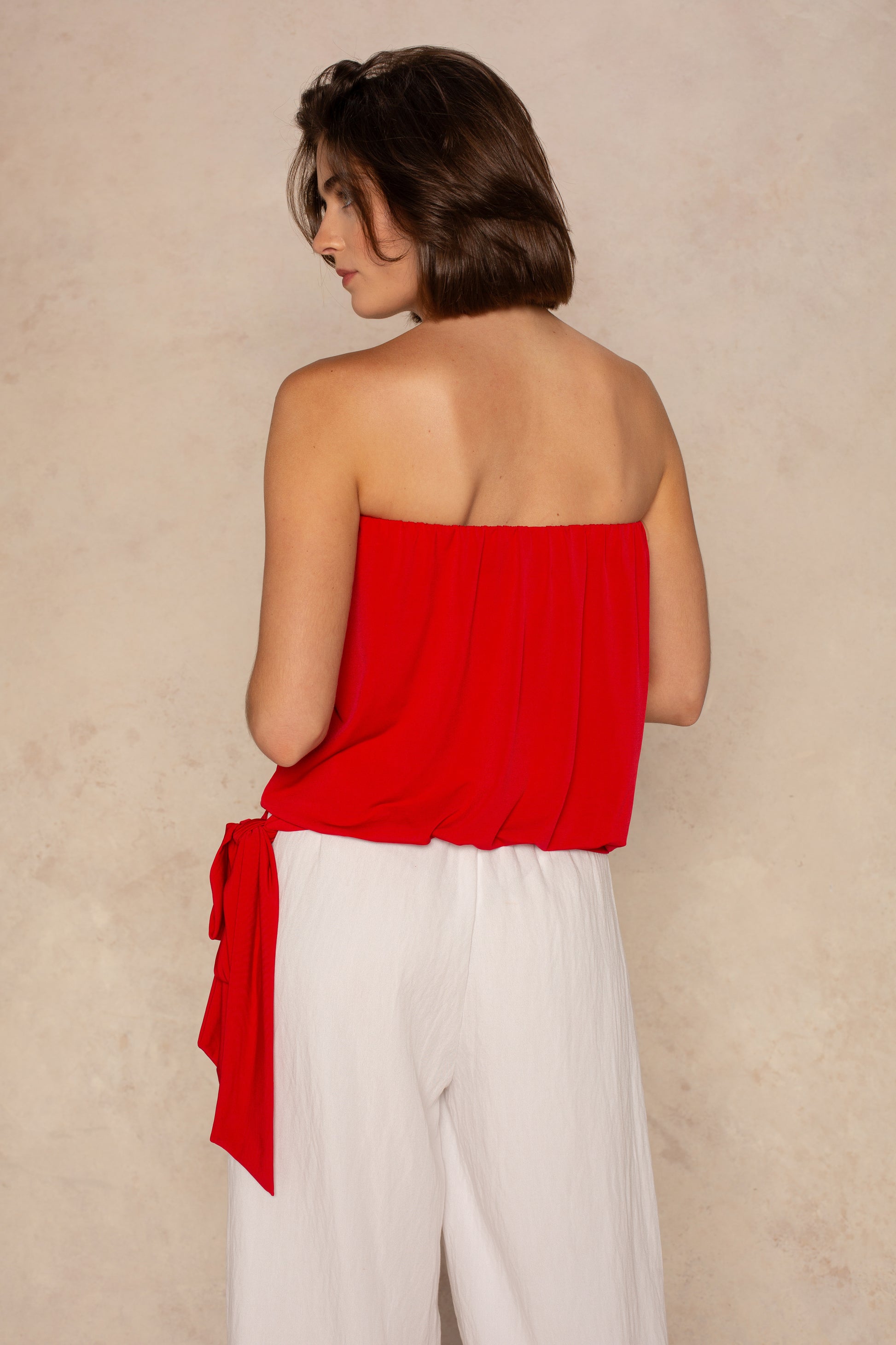 A woman with short brown hair faces away, wearing the Georgie Gemma Side Tie Tube Top and high-waisted, loose white pants—a chic summer outfit against a light beige background.