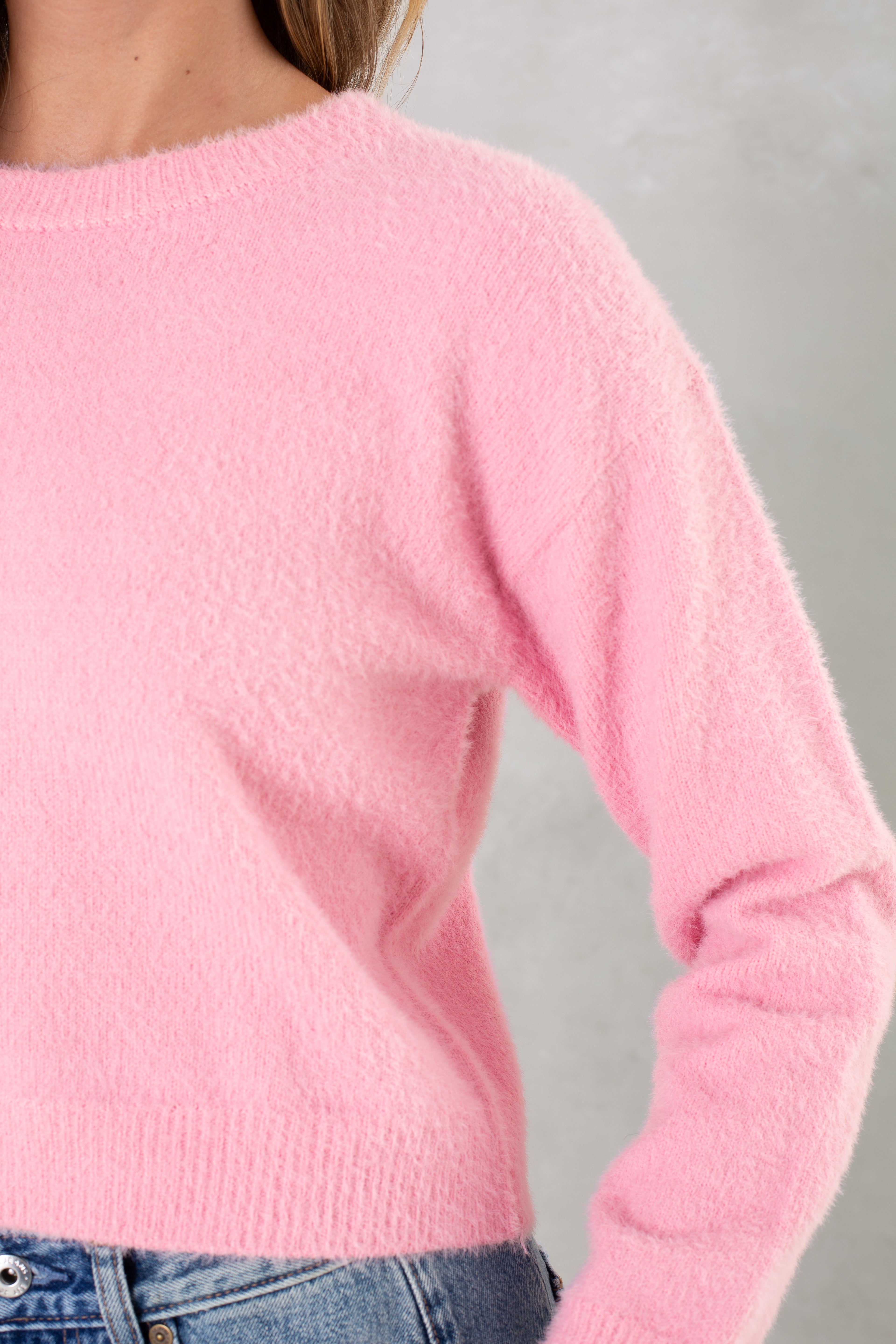 A person models the Isla Eyelash Sweater in soft pink with light blue denim jeans, shown from shoulders to hips against a neutral background. The face is not visible.
