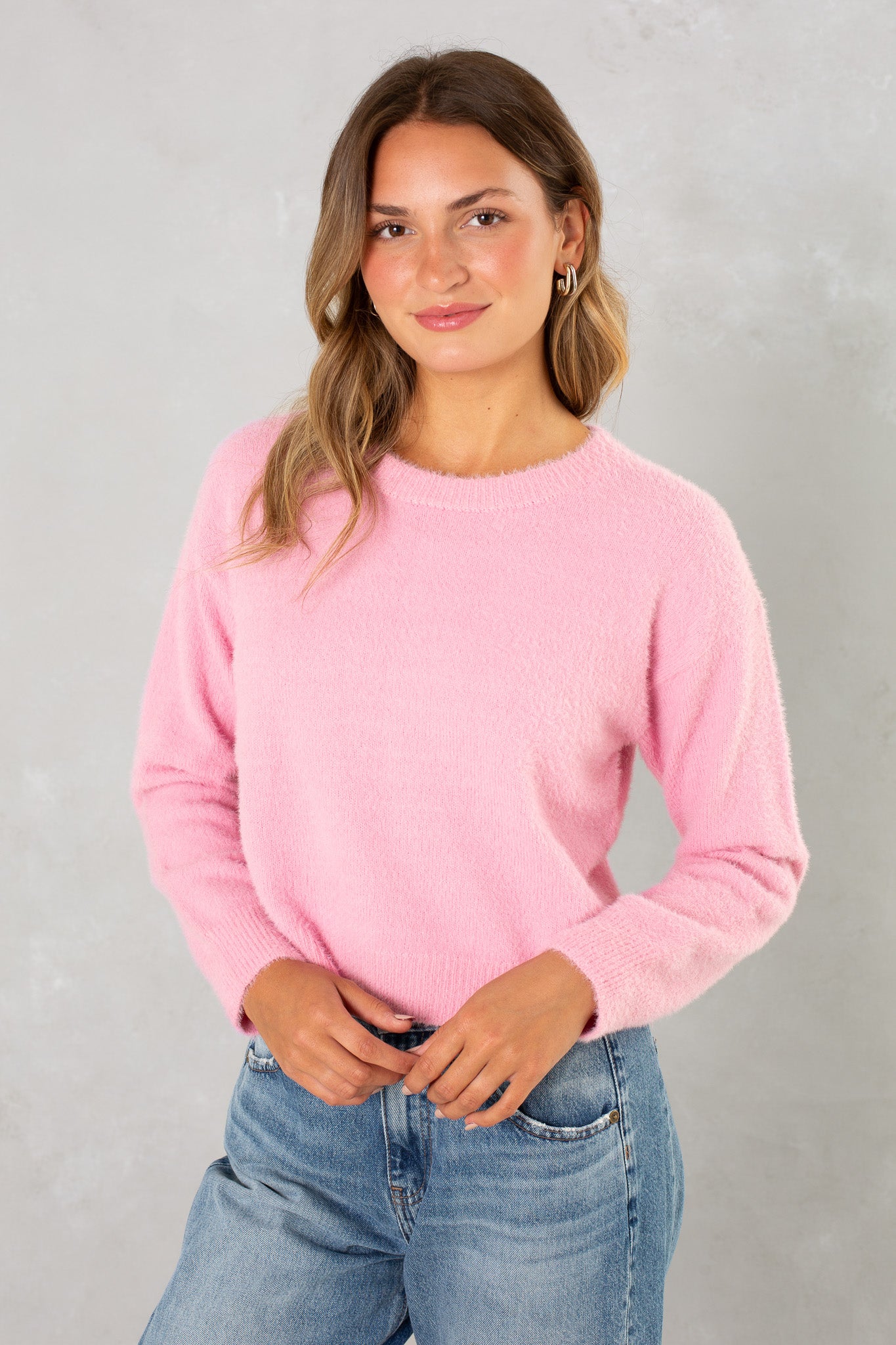 A woman with wavy brown hair wears Isla’s Eyelash Sweater in light pink and blue jeans, standing against a light gray background and smiling gently with her hands in front of her.