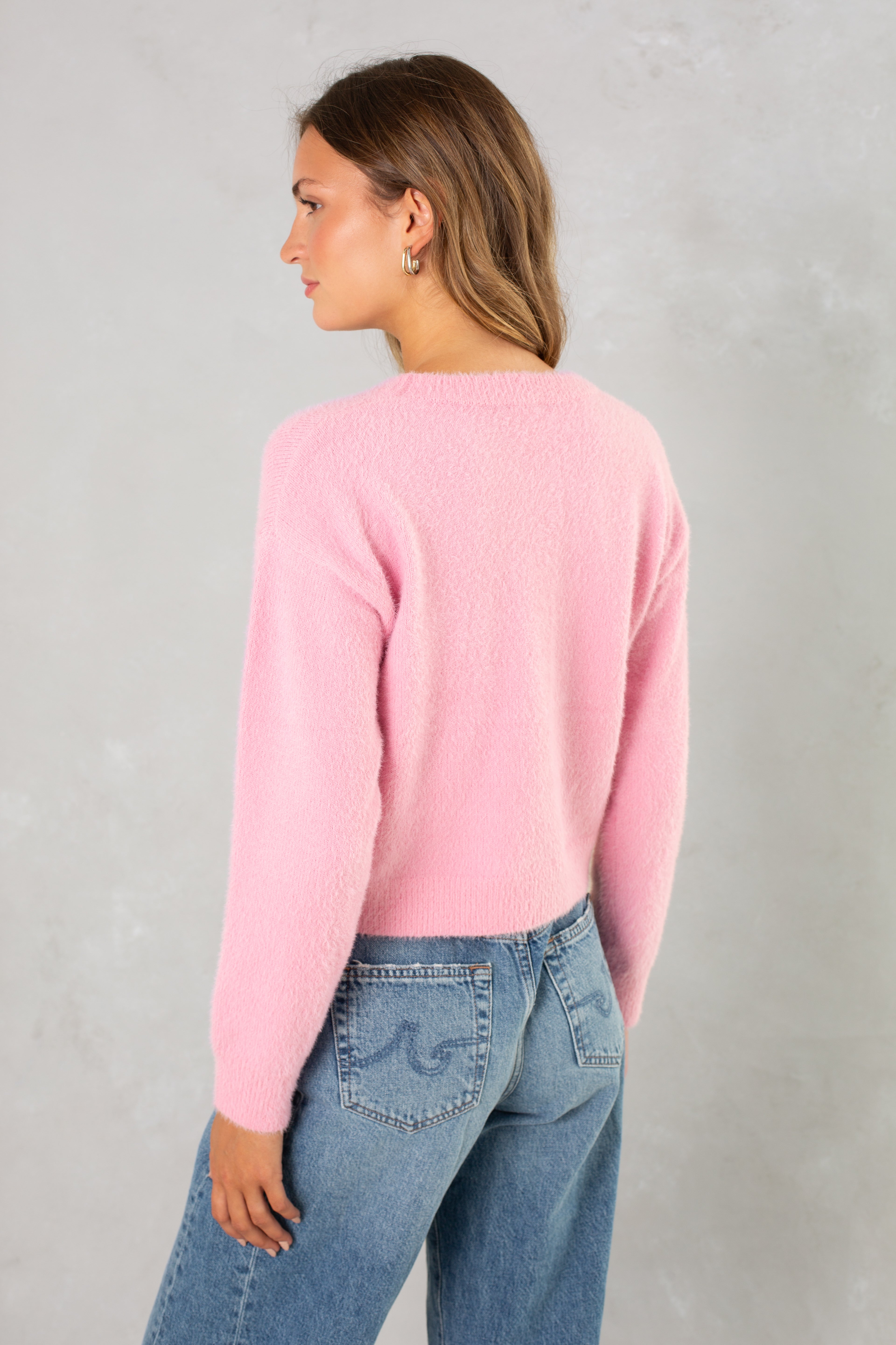 A woman with long brown hair wears the Isla Eyelash Sweater in light pink, paired with blue jeans, and stands with her back to the camera against a light gray background.
