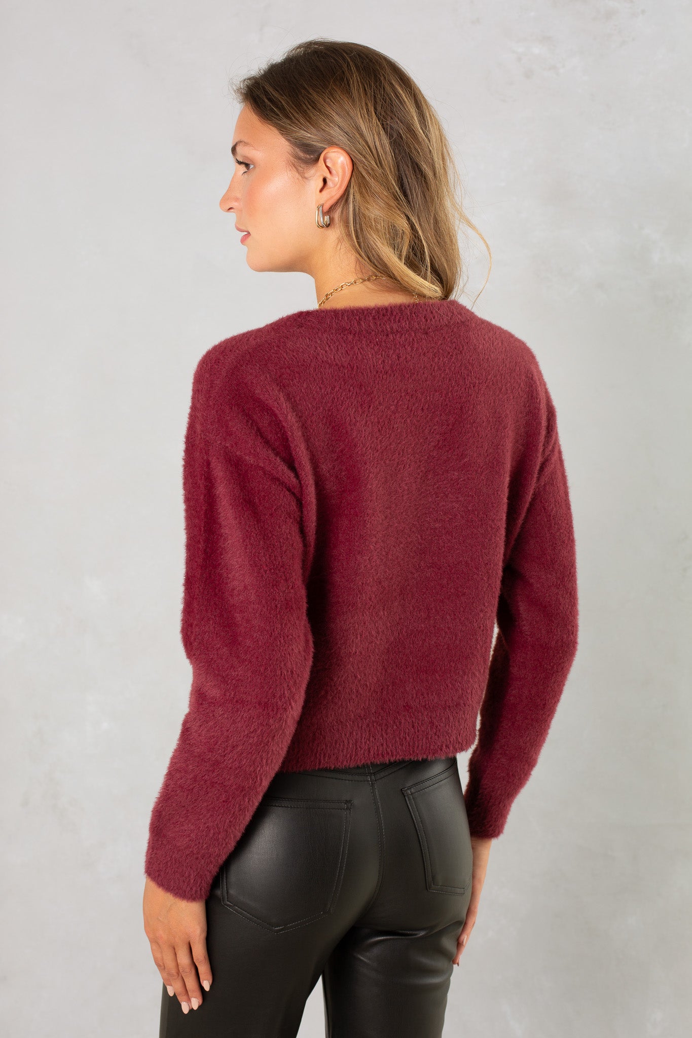 A woman with wavy light brown hair is seen from the back wearing Isla's Eyelash Sweater in maroon and high-waisted black leather pants against a light grey background.