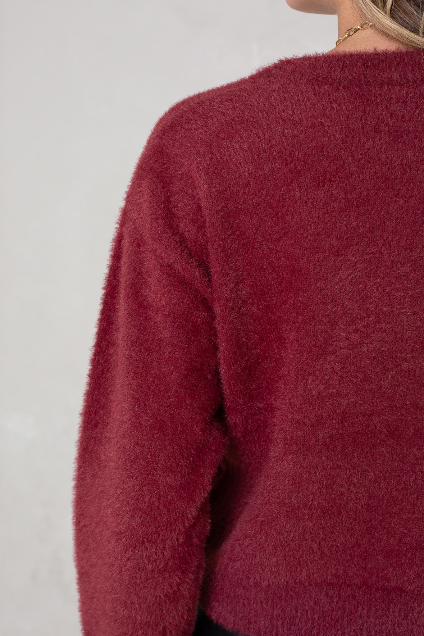 Close-up of a person wearing the Isla Eyelash Sweater in dark red. The image shows the soft, fuzzy texture on the back and shoulder, with part of blonde hair and a necklace visible against a plain, light background.