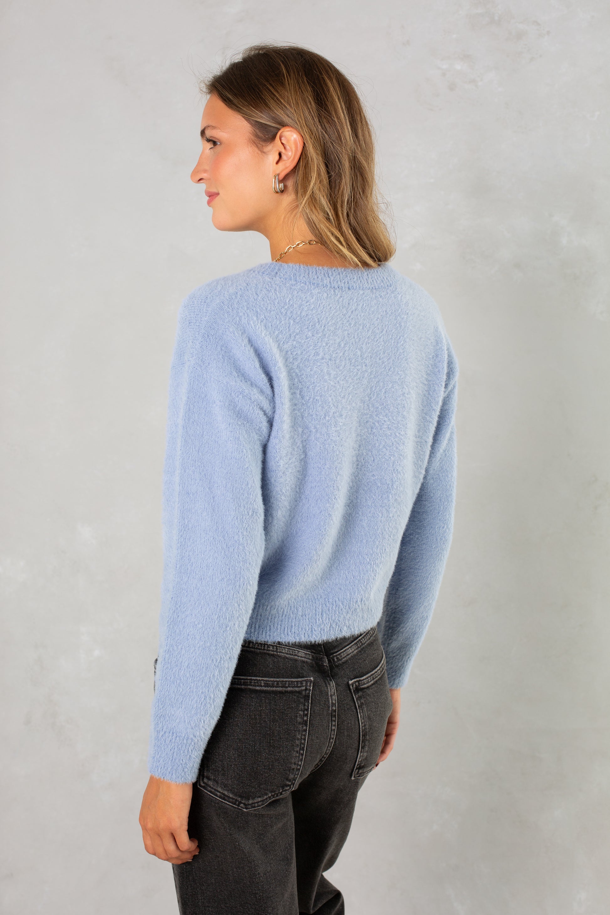 A woman with light brown hair stands facing away, looking left, wearing Isla's Eyelash Sweater in light blue and high-waisted black jeans against a light gray textured background.
