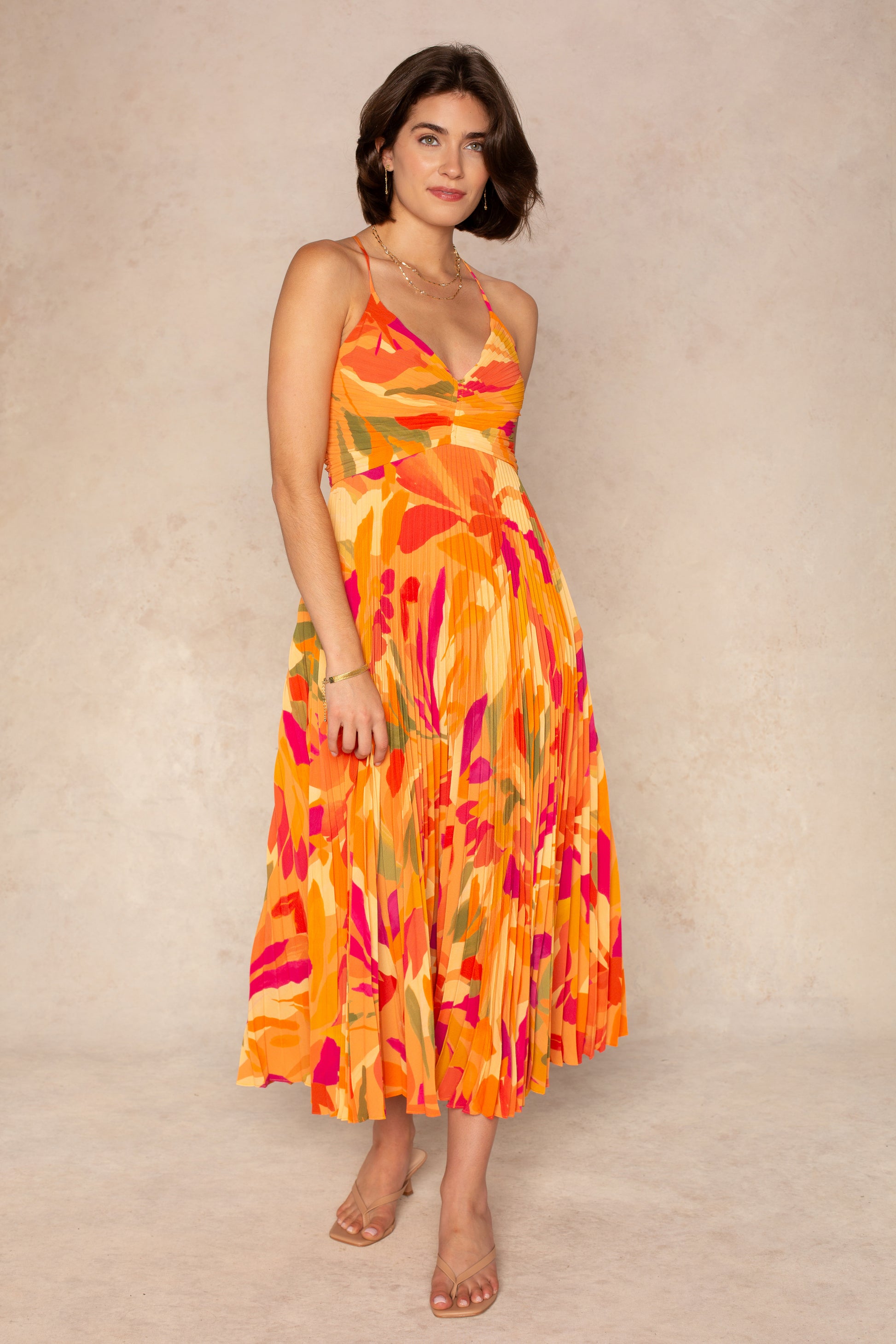 A woman wears the ASTR Blythe Midi Dress in Orange Multi, a sleeveless plissé style with adjustable spaghetti straps and an abstract floral pattern, paired with clear-strap sandals and gold jewelry against a neutral background.