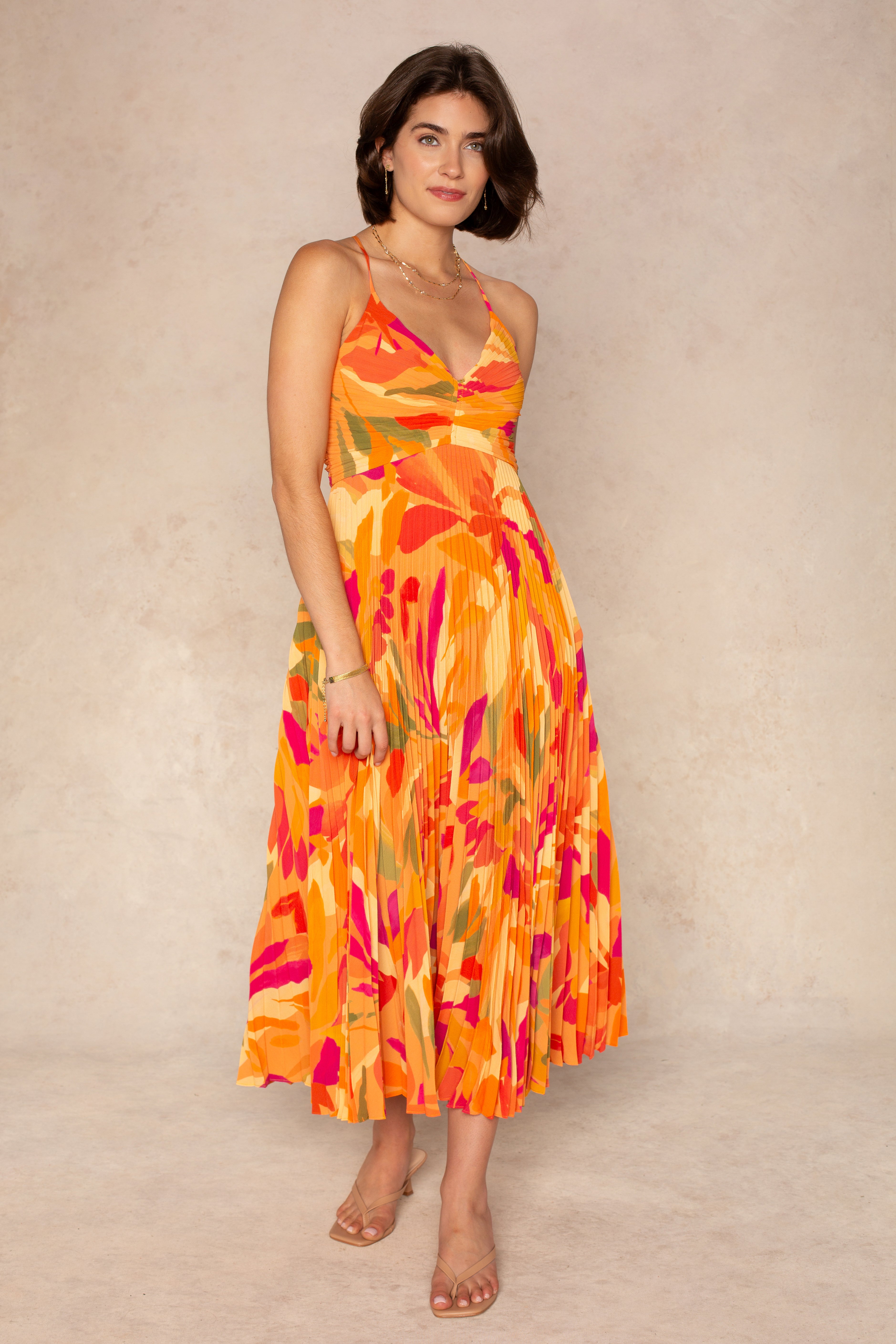 A woman wears the ASTR Blythe Midi Dress in Orange Multi, a sleeveless plissé style with adjustable spaghetti straps and an abstract floral pattern, paired with clear-strap sandals and gold jewelry against a neutral background.
