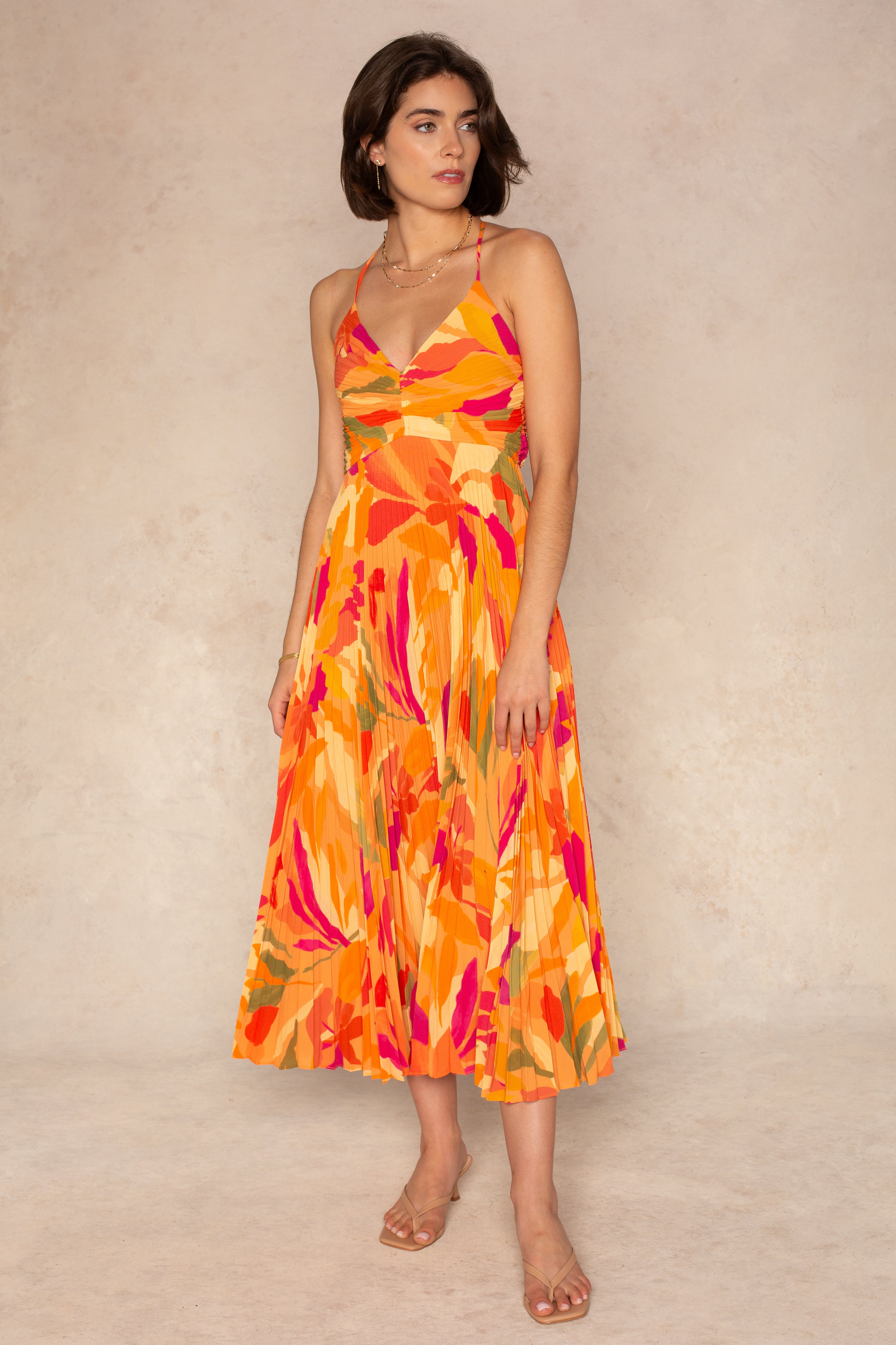A woman stands on a light background wearing the Blythe Midi Dress by ASTR, featuring a V-neck, orange and pink floral print, pleated skirt, and adjustable spaghetti straps, paired with clear-heeled sandals and layered necklaces.