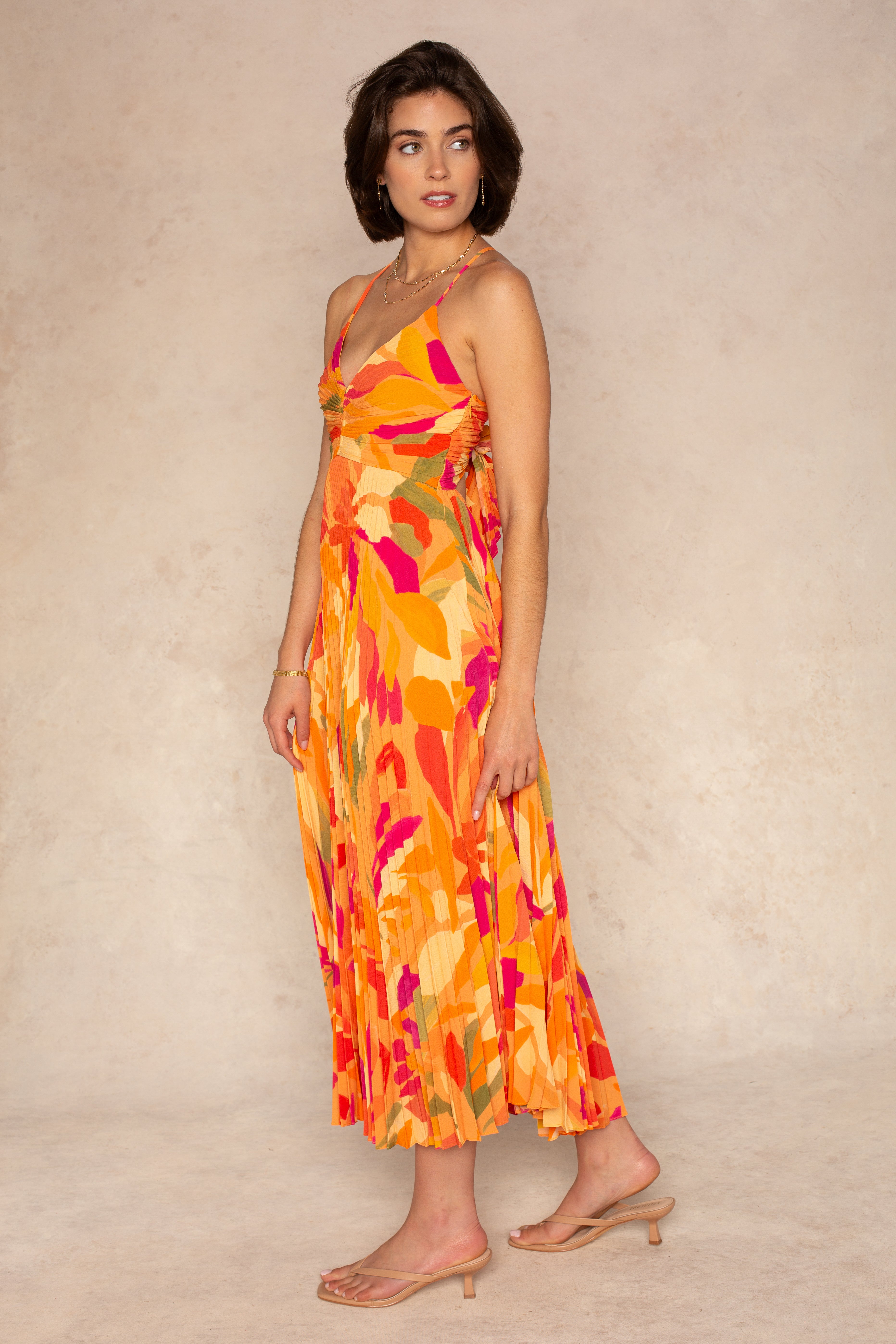 A woman models the ASTR Blythe Midi Dress, featuring adjustable spaghetti straps and a pleated orange and pink floral skirt. She pairs it with nude heeled sandals, standing against a neutral background, gazing left with one hand by her side.