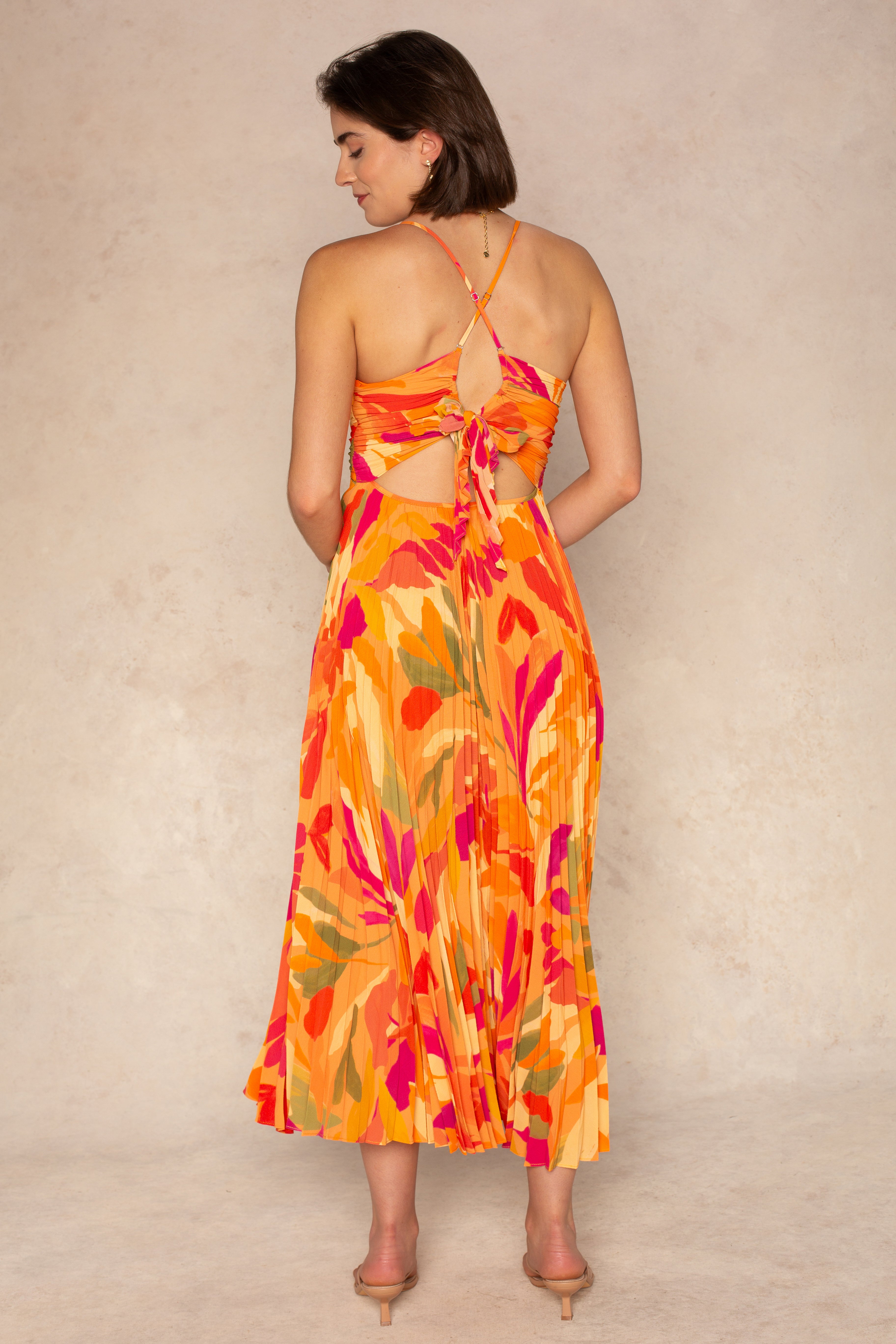 A woman with short brown hair and nude heels stands against a beige background, wearing the ASTR Blythe Midi Dress—a sleeveless, backless orange and red floral plissé dress with adjustable spaghetti straps and a pleated skirt.