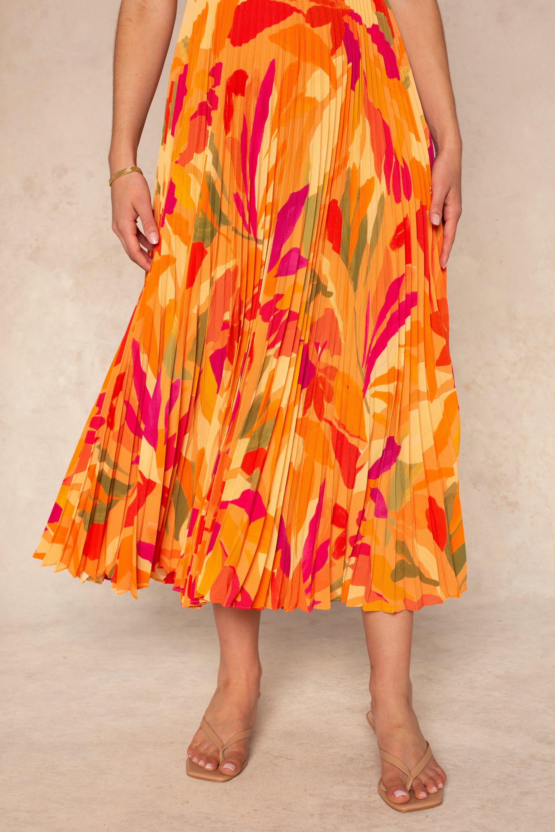 A woman wears the ASTR Blythe Midi Dress, featuring a vibrant orange, red, pink, and yellow leaf print with playful pleats, paired with strappy nude sandals against a neutral background.