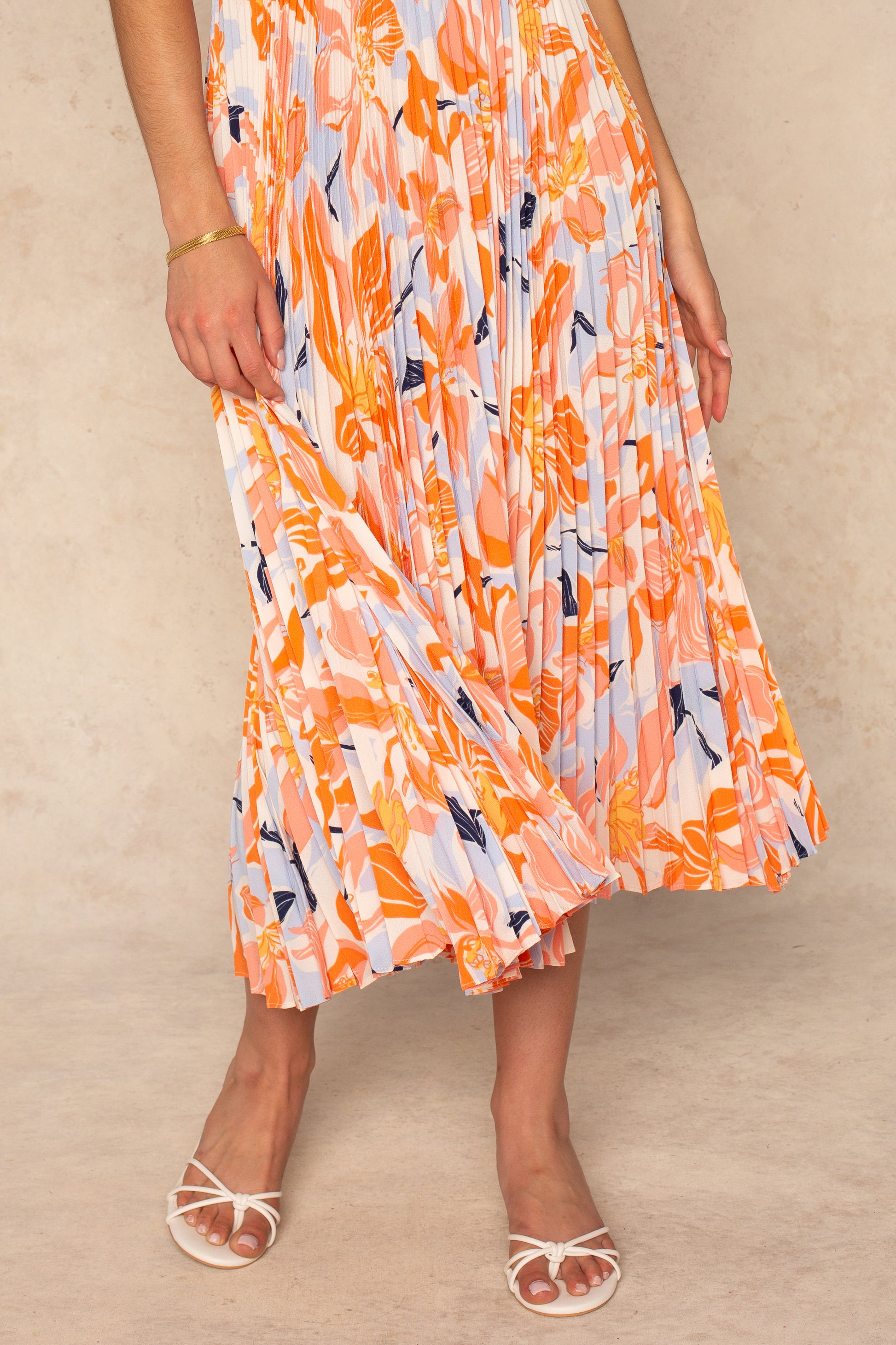 A person wearing the ASTR Blythe Midi Dress in orange, white, and blue stands on a light surface, holding the skirt. They pair it with white strappy heeled sandals and a gold bracelet. Their face is not visible.