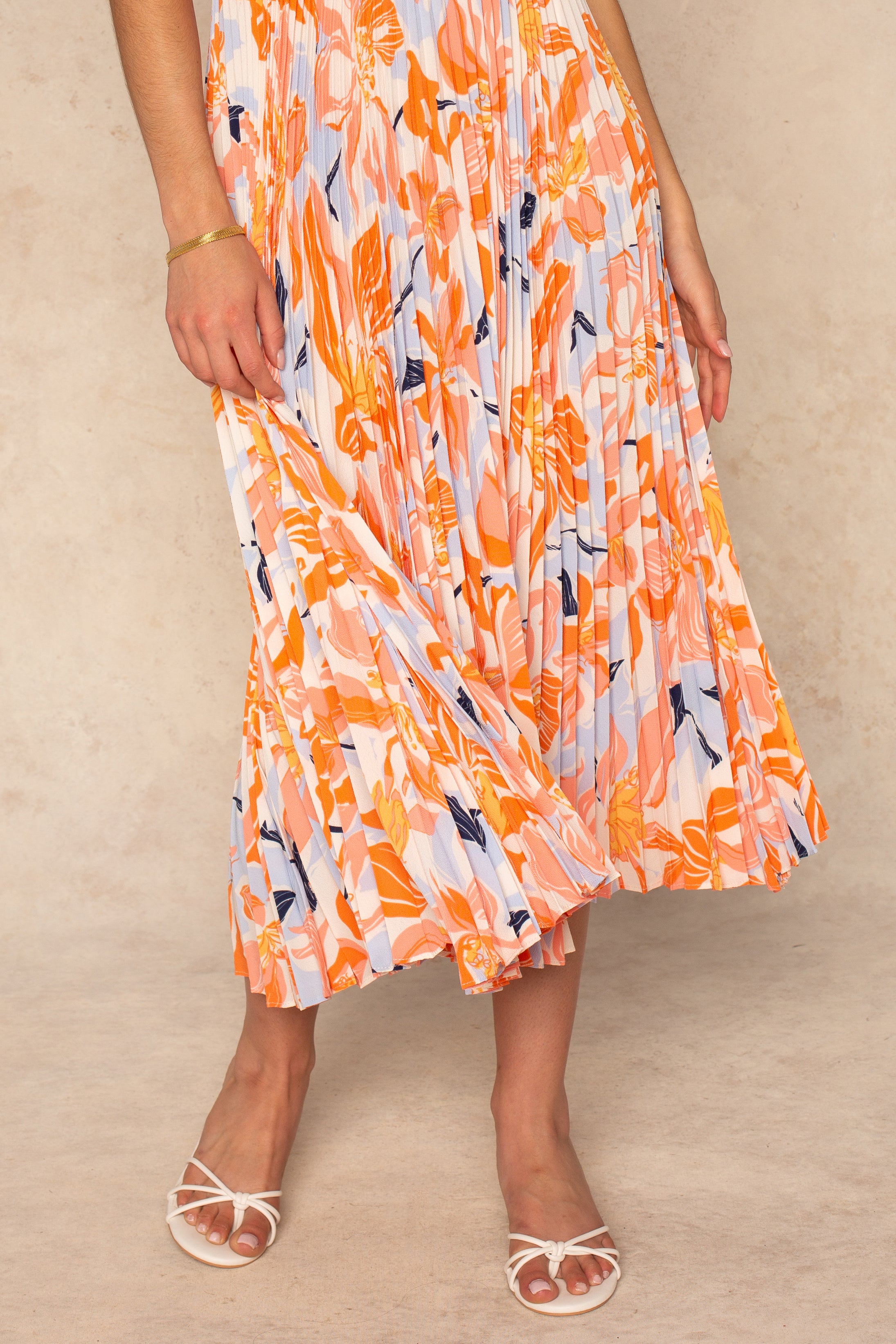 A person wearing the ASTR Blythe Midi Dress in orange, white, and blue stands on a light surface, holding the skirt. They pair it with white strappy heeled sandals and a gold bracelet. Their face is not visible.