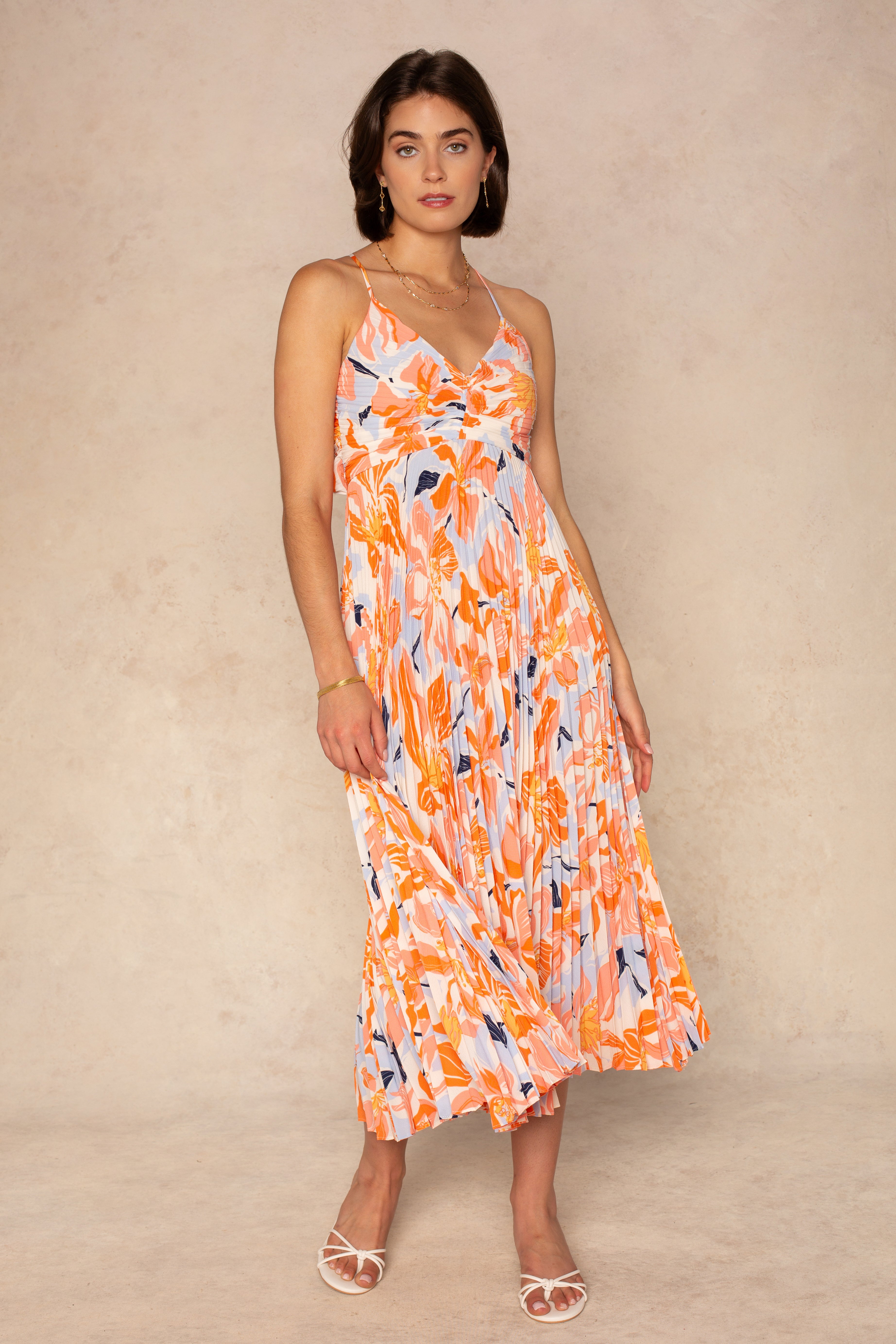 A woman stands against a beige background wearing the ASTR Blythe Midi Dress, a sleeveless orange and blue plissé midi with adjustable spaghetti straps. She has shoulder-length brown hair and white open-toed heels.