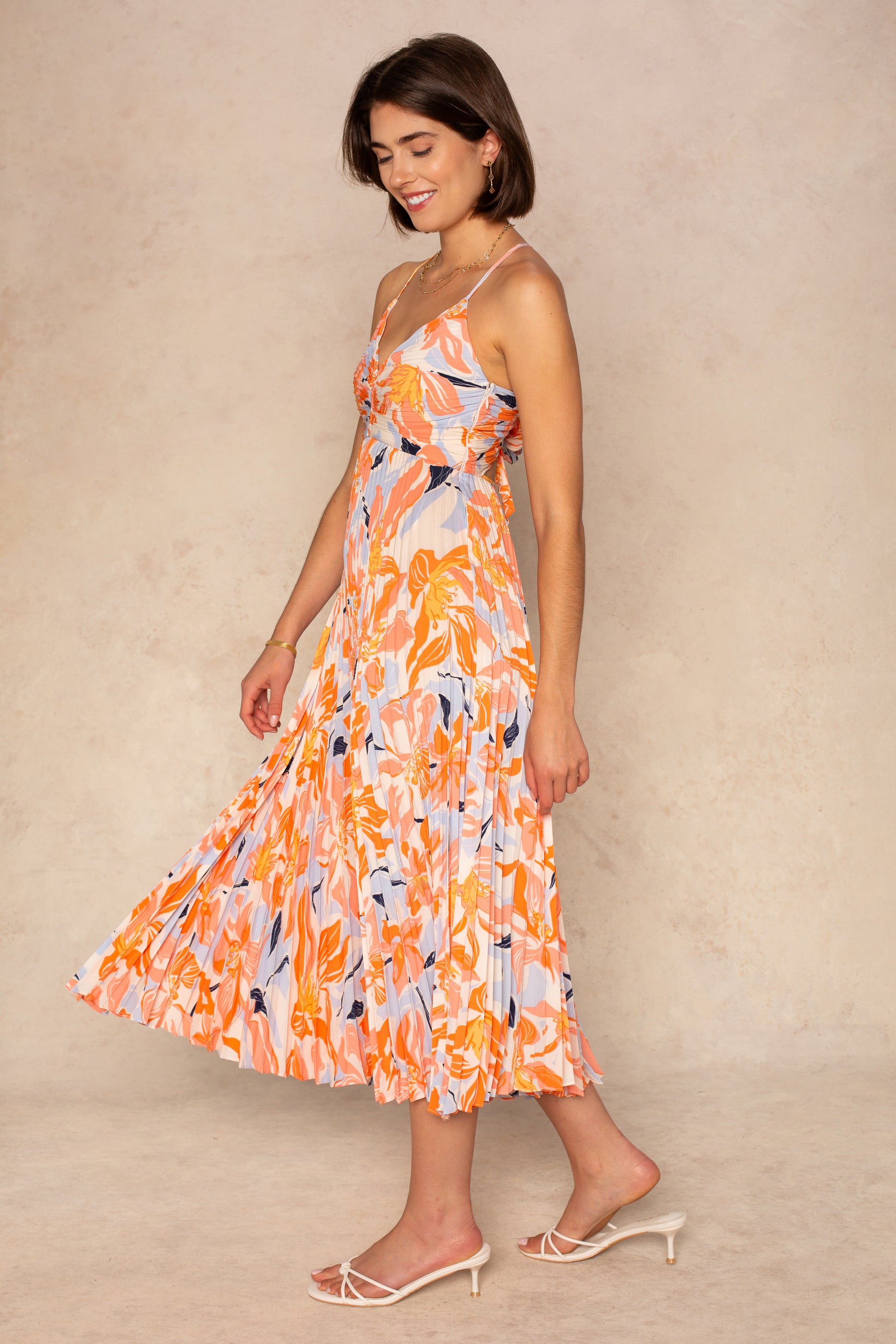 A woman smiles, looking down, in the ASTR Blythe Midi Dress—a sleeveless orange and white plissé midi with a pleated skirt—paired with white heeled sandals against a beige background.