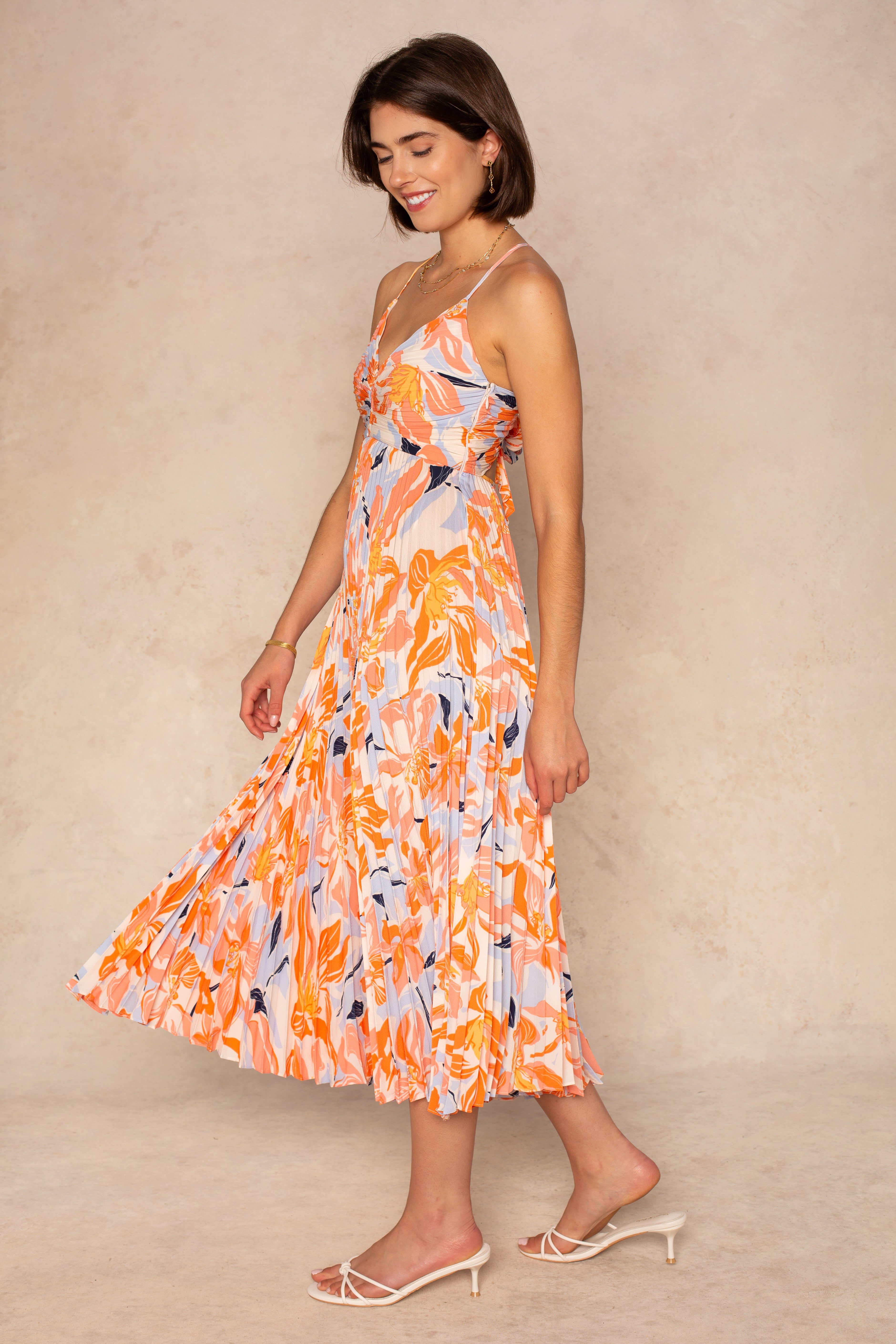 A woman smiles, looking down, in the ASTR Blythe Midi Dress—a sleeveless orange and white plissé midi with a pleated skirt—paired with white heeled sandals against a beige background.