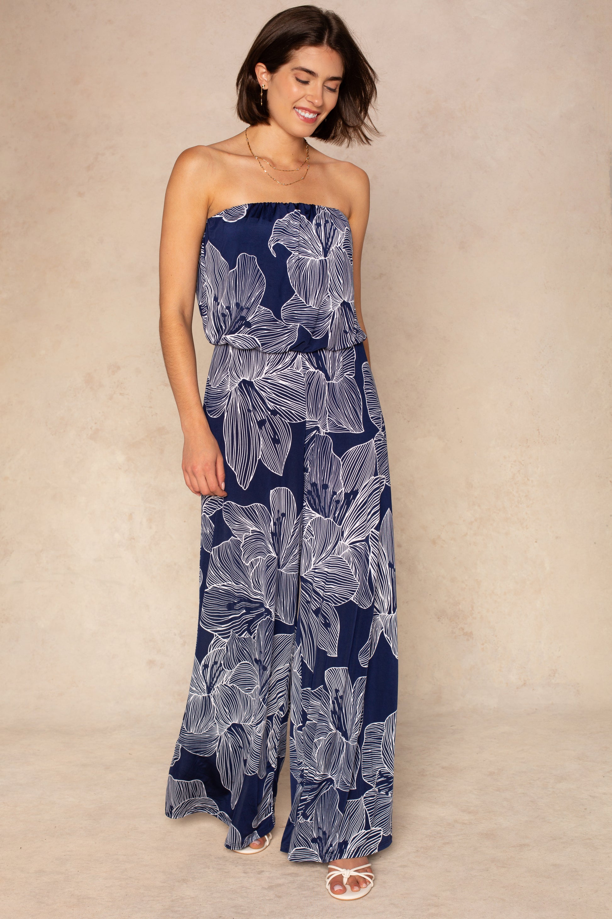 A woman smiles against a beige background, wearing the Isla Ari Strapless Easy Jumpsuit with a navy floral print and wide-leg pants, paired with white sandals.