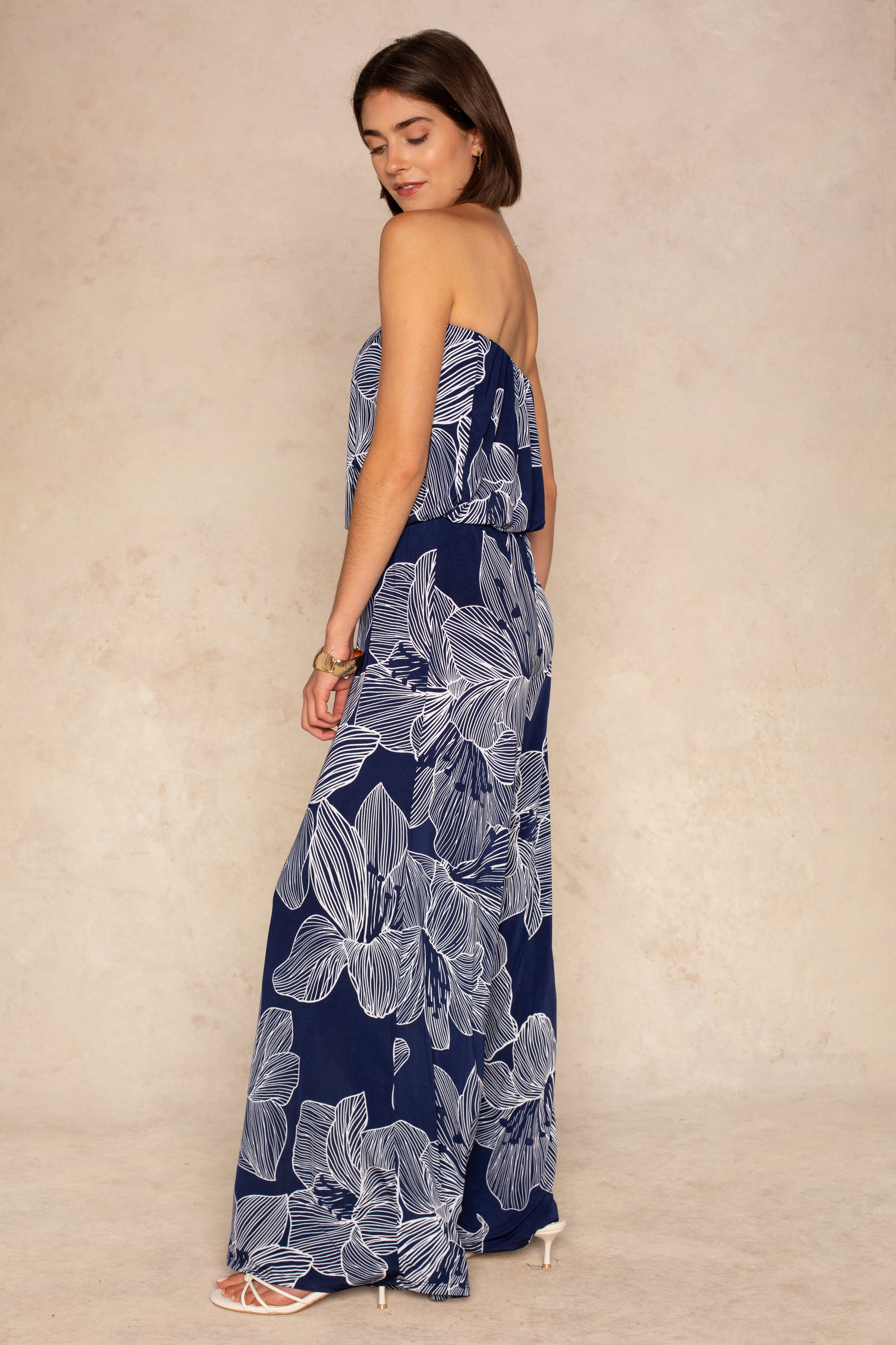 A woman wears the Isla Ari Strapless Easy Jumpsuit, featuring a navy floral print and wide-leg pants. She stands sideways, looking over her shoulder, paired with white heeled sandals and a gold bracelet against a neutral background.