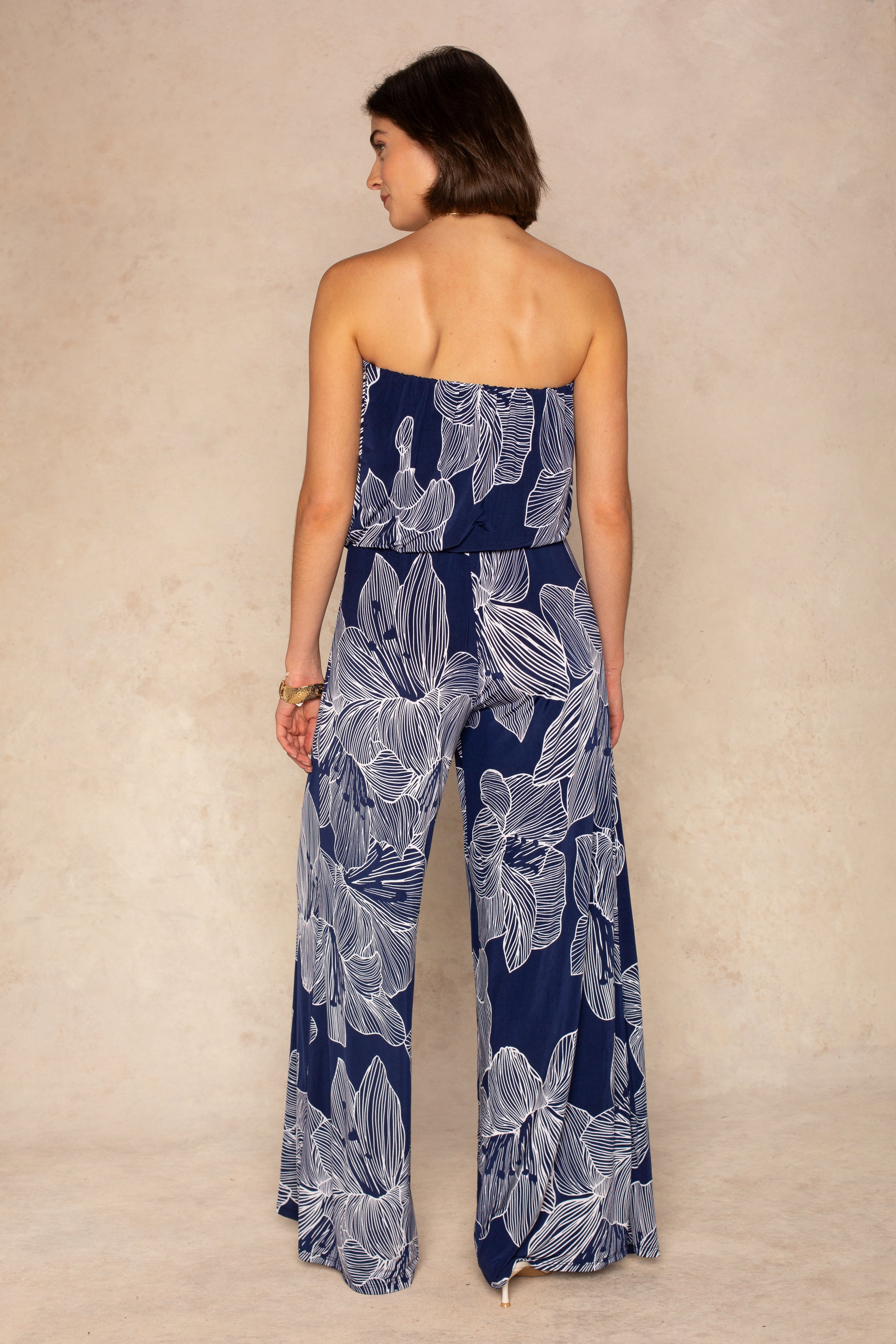 A woman with short brown hair poses against a light beige background, wearing the Isla Ari Strapless Easy Jumpsuit in a navy floral print with wide-leg pants.