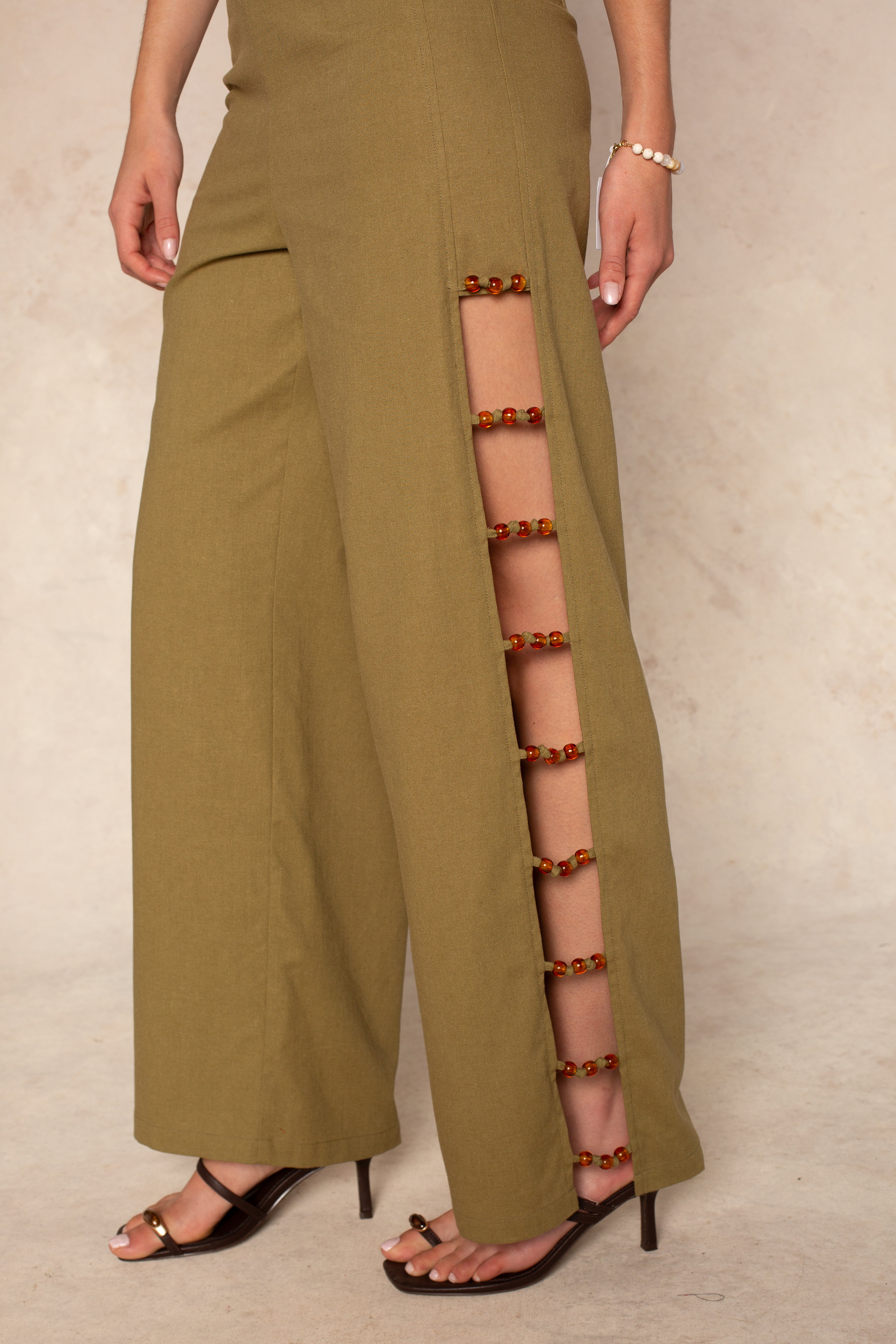 A woman in the Georgie Harper Linen Beaded Pant—boho olive green wide-leg pants with red and gold beaded side slits—styled with brown heeled sandals and a beaded bracelet for a chic, resort-ready look.
