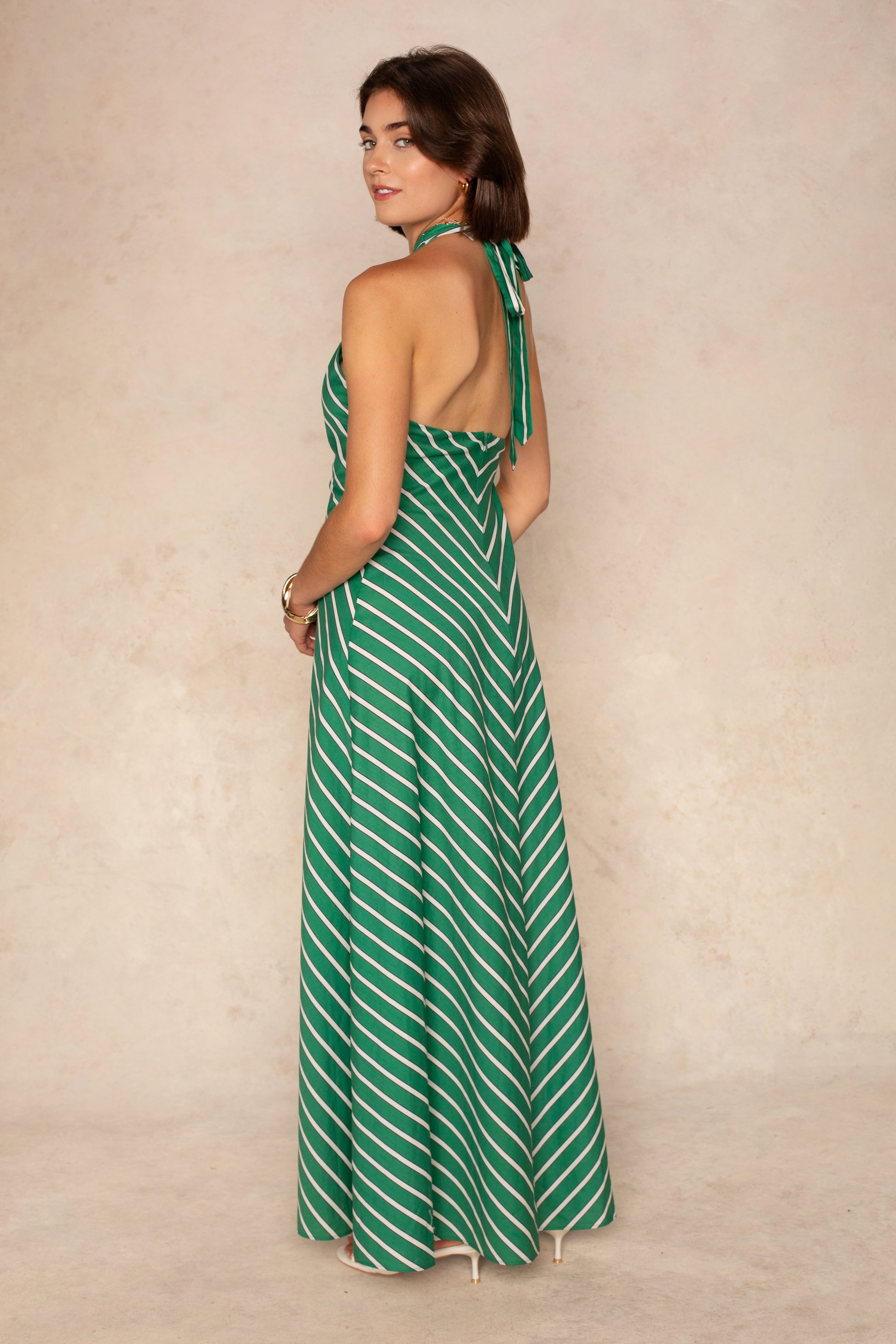 A woman poses with her back to the camera in the Georgie Parker Halter Maxi Dress, featuring green and white stripes. The floor-length summer dress is styled with white heels against a beige backdrop.