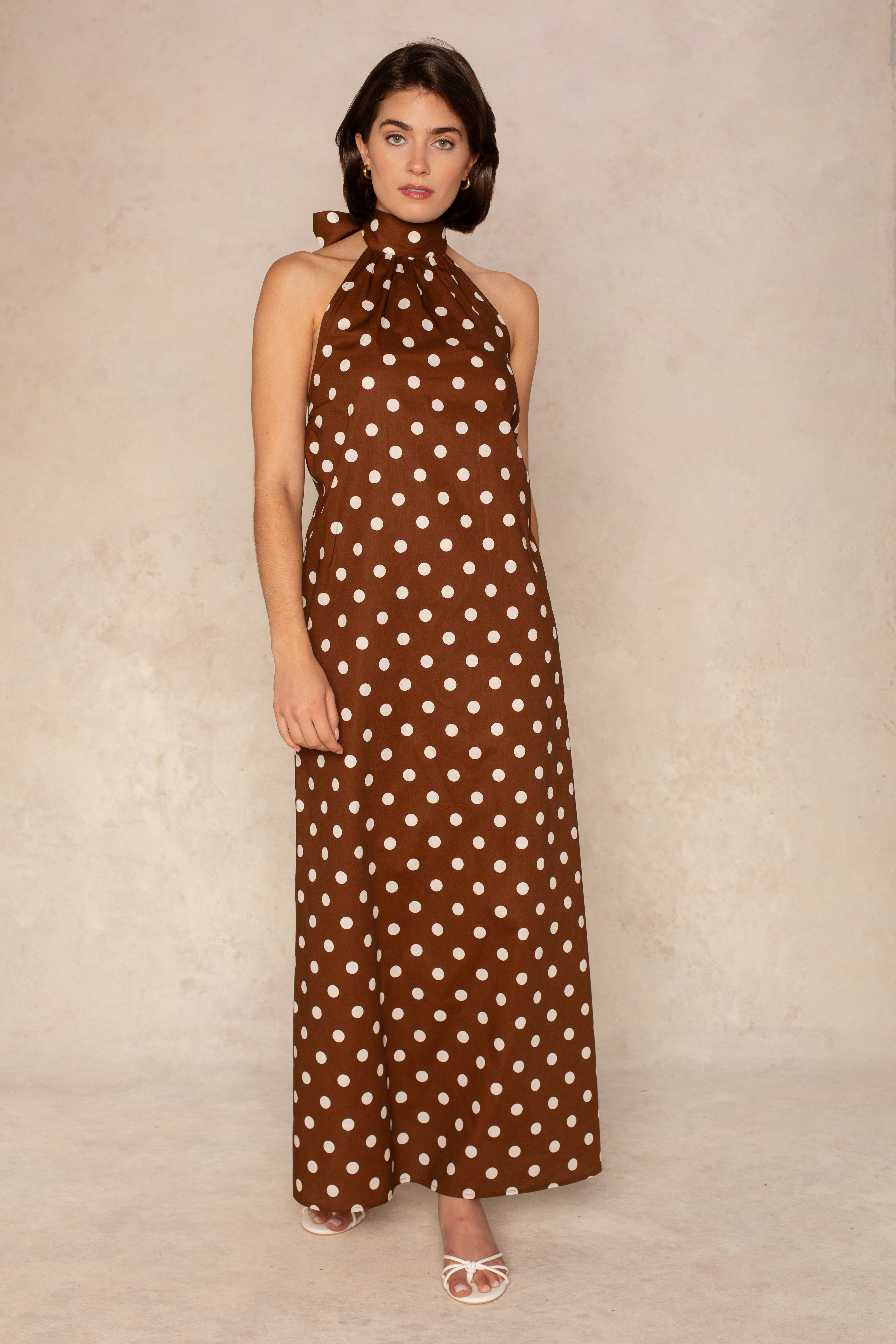 A woman with short dark hair and a neutral expression models the Georgie Laney Halter Maxi Dress, a sleeveless brown polka dot piece, against a neutral background. She pairs it with white open-toe heels.