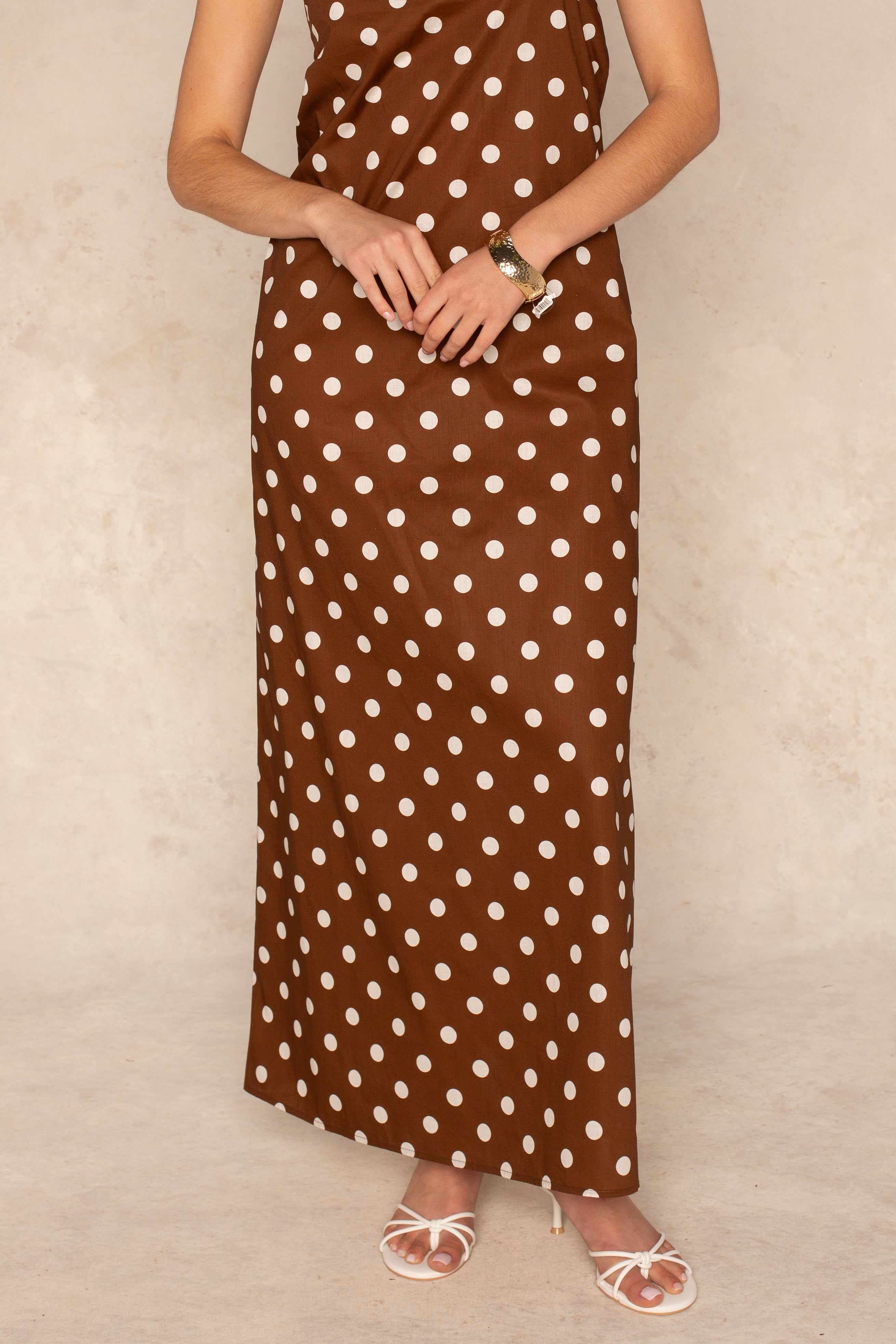 A person models the Georgie Laney Halter Maxi Dress, featuring white polka dots, styled with white strappy heels and a bracelet against a neutral background. The head is not visible in the image.