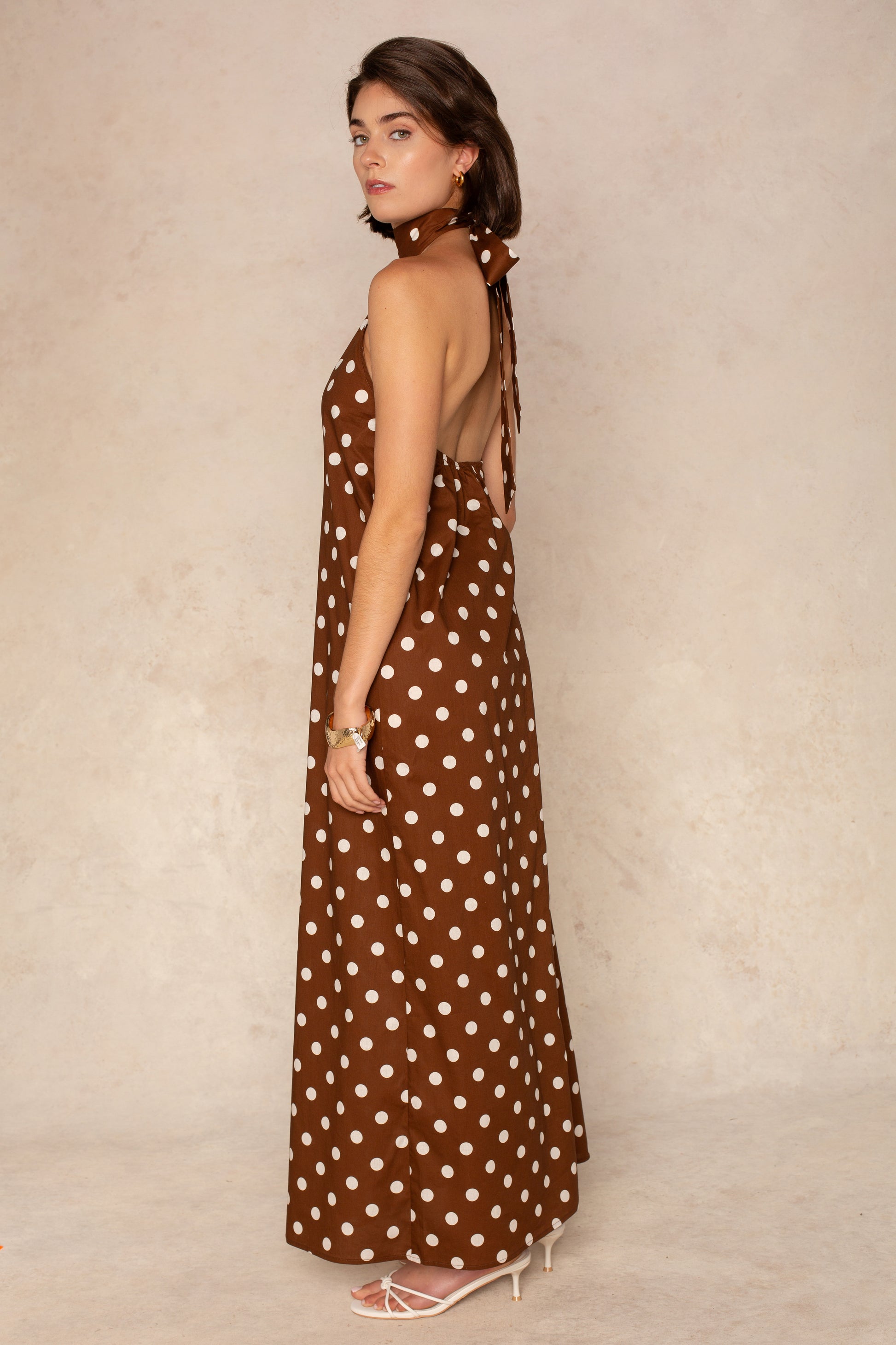 A woman stands sideways in the Georgie Laney Halter Maxi Dress, a brown polka dot maxi with an open back, paired with white heeled sandals against a neutral beige background.