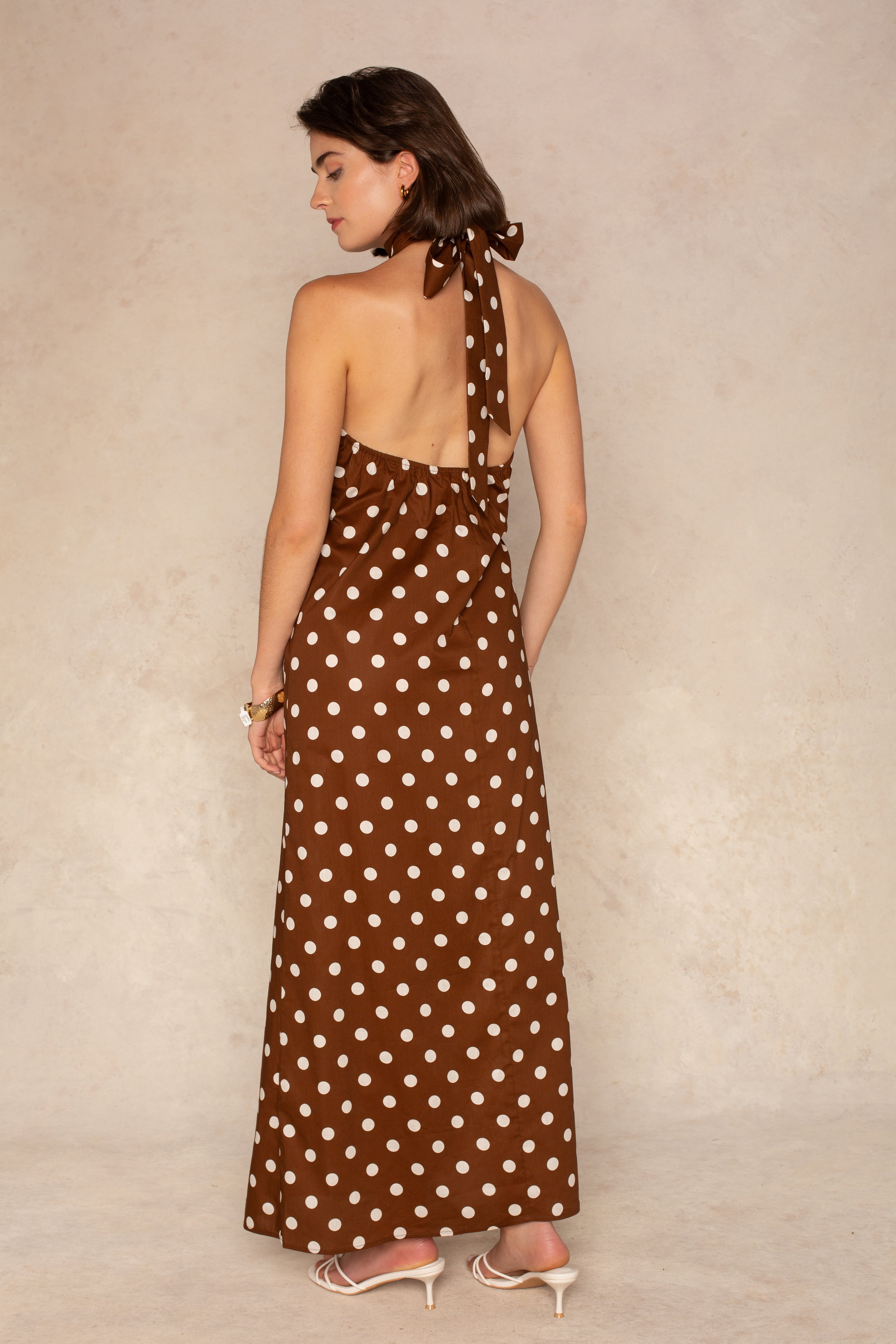 A woman with short brown hair wears the Georgie Laney Halter Maxi Dress, a brown maxi dress with white polka dots and an open back. She pairs it with white heeled sandals and stands against a neutral beige background.