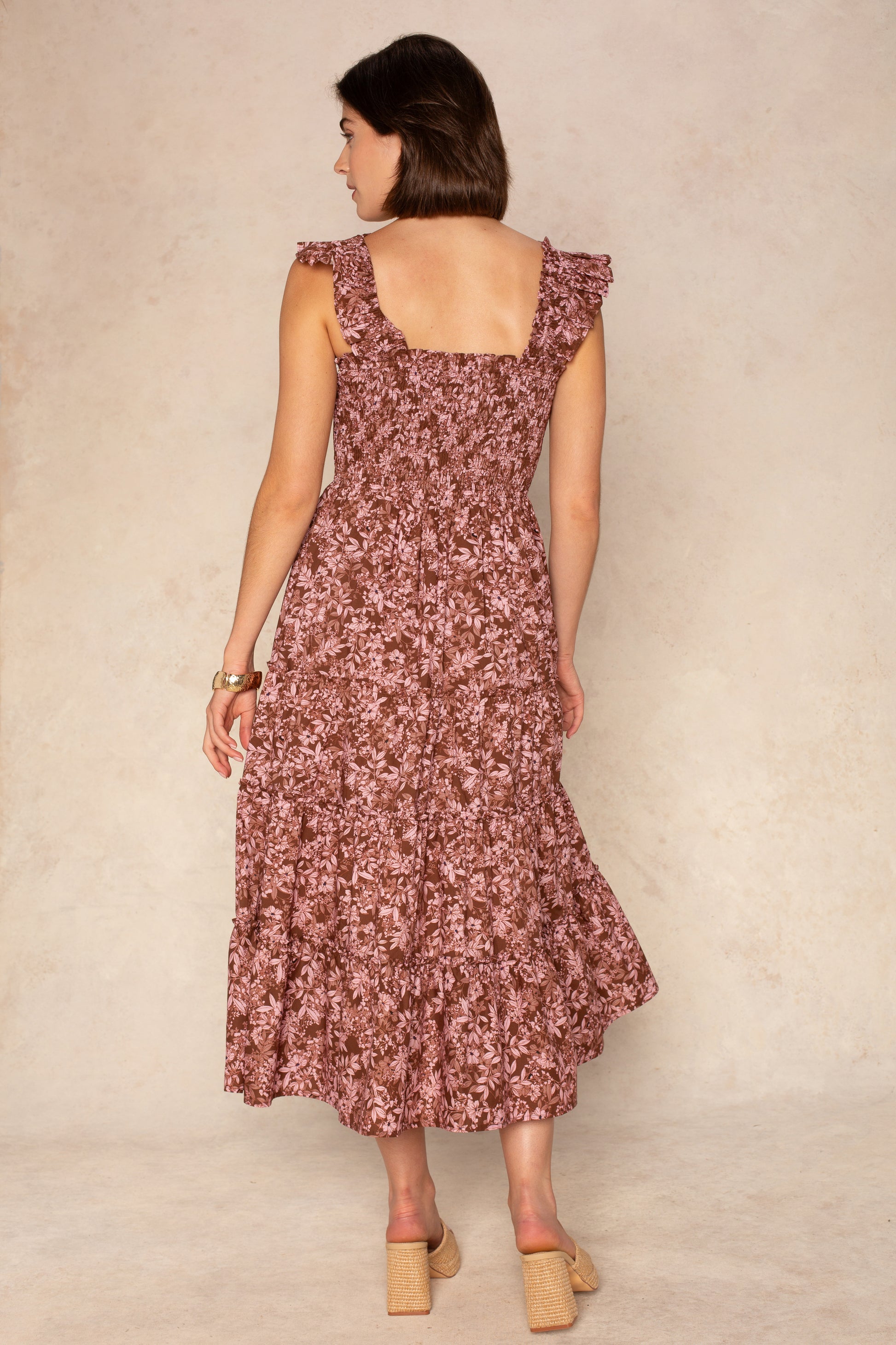 A woman with short brown hair models the Georgie Calypso Smocked Midi Dress, a sleeveless mauve floral piece with a tiered skirt and ruffles. She pairs it with beige block-heeled shoes against a light neutral background.