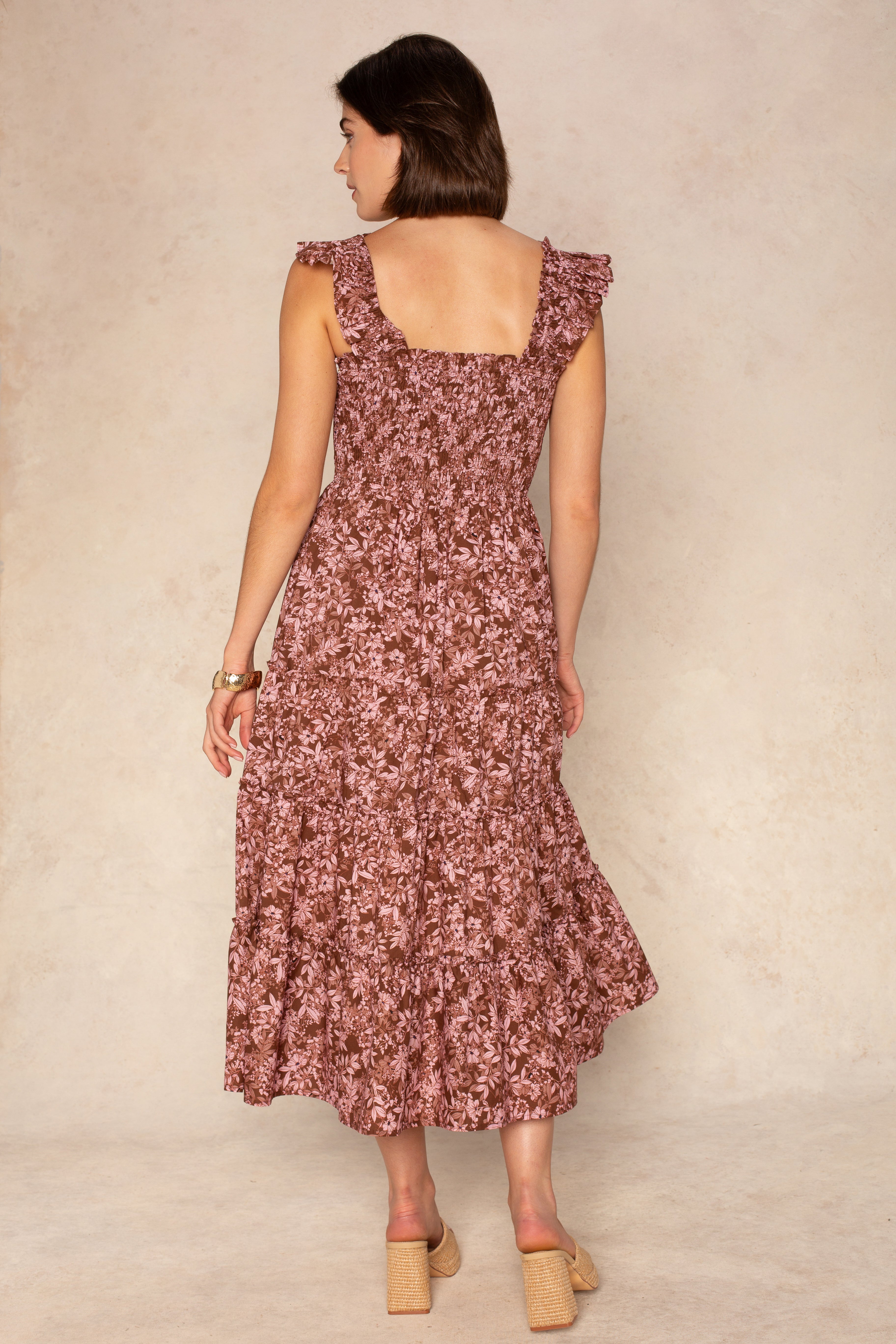 A woman with short brown hair models the Georgie Calypso Smocked Midi Dress, a sleeveless mauve floral piece with a tiered skirt and ruffles. She pairs it with beige block-heeled shoes against a light neutral background.